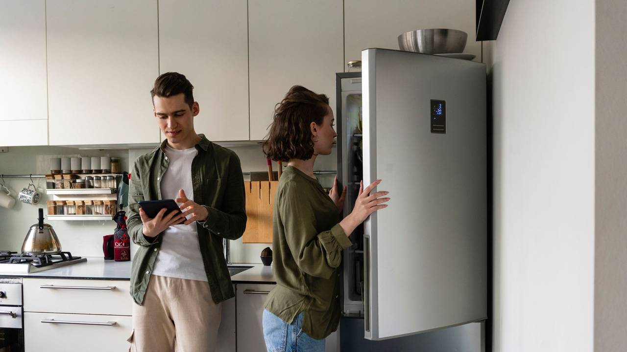 Auf dem Foto sieht man eine Frau einen Kühlschrank öffnen, neben ihr steht ein Mann, welcher auf sein Tablet schaut.