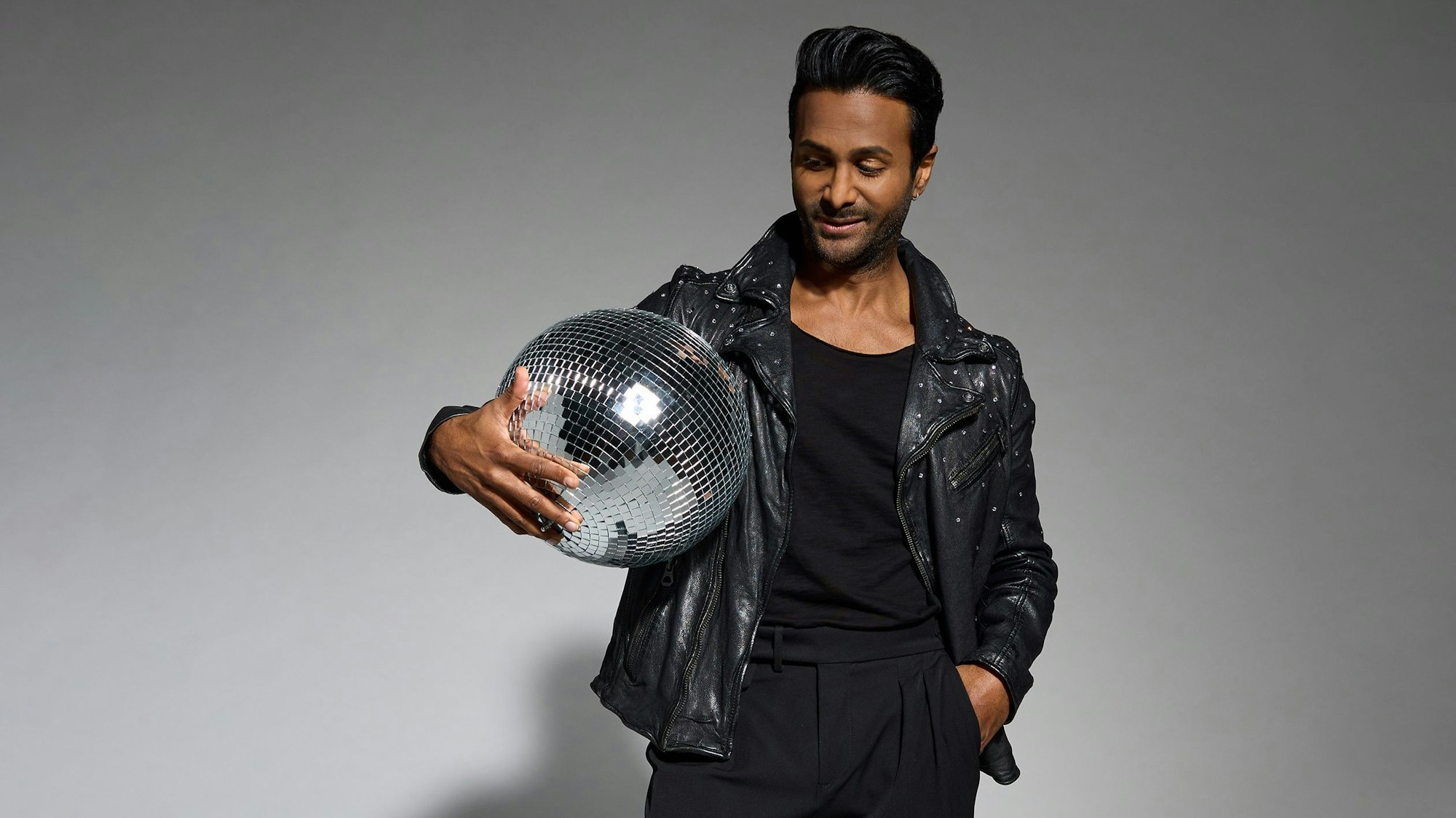 Biyon Kattilathu posiert für die Kamera. Er trägt eine schwarze Hose und eine schwarze Lederjacke, im Arm hält er eine Diskokugel.