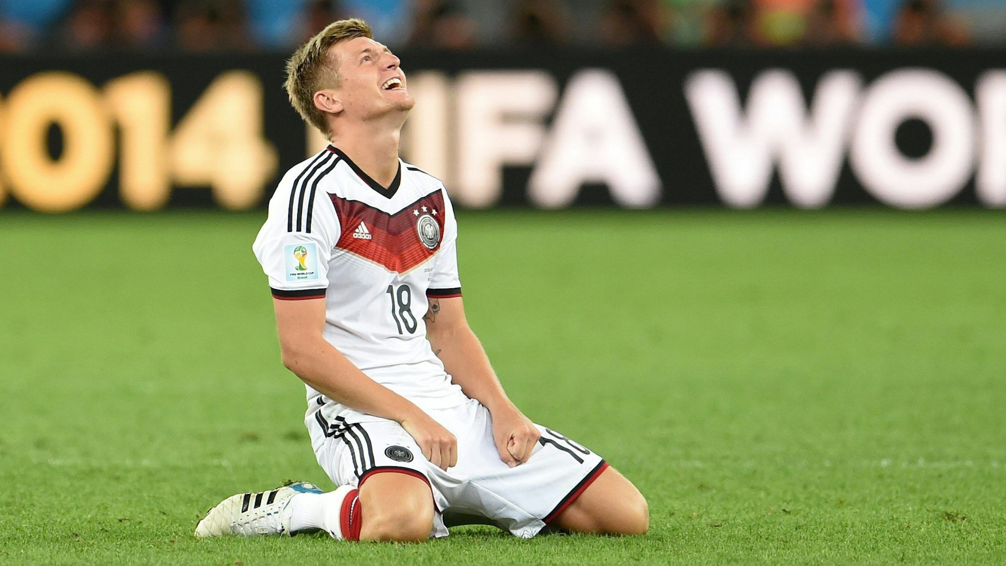 Toni Kroos kniet auf dem Rasen und guckt lachend nach oben.