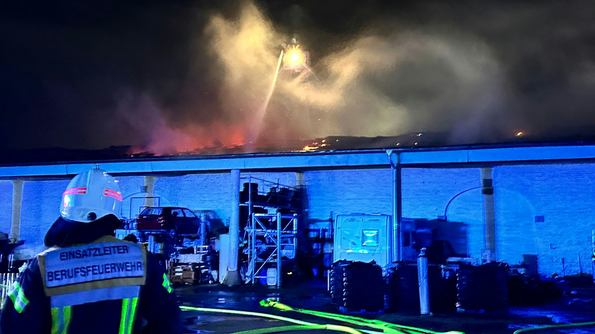 Ein Einsatzleiter der Feuerwehr steht vor einem brennenden Gebäude und beobachtet die Arbeit seiner Kollegen.