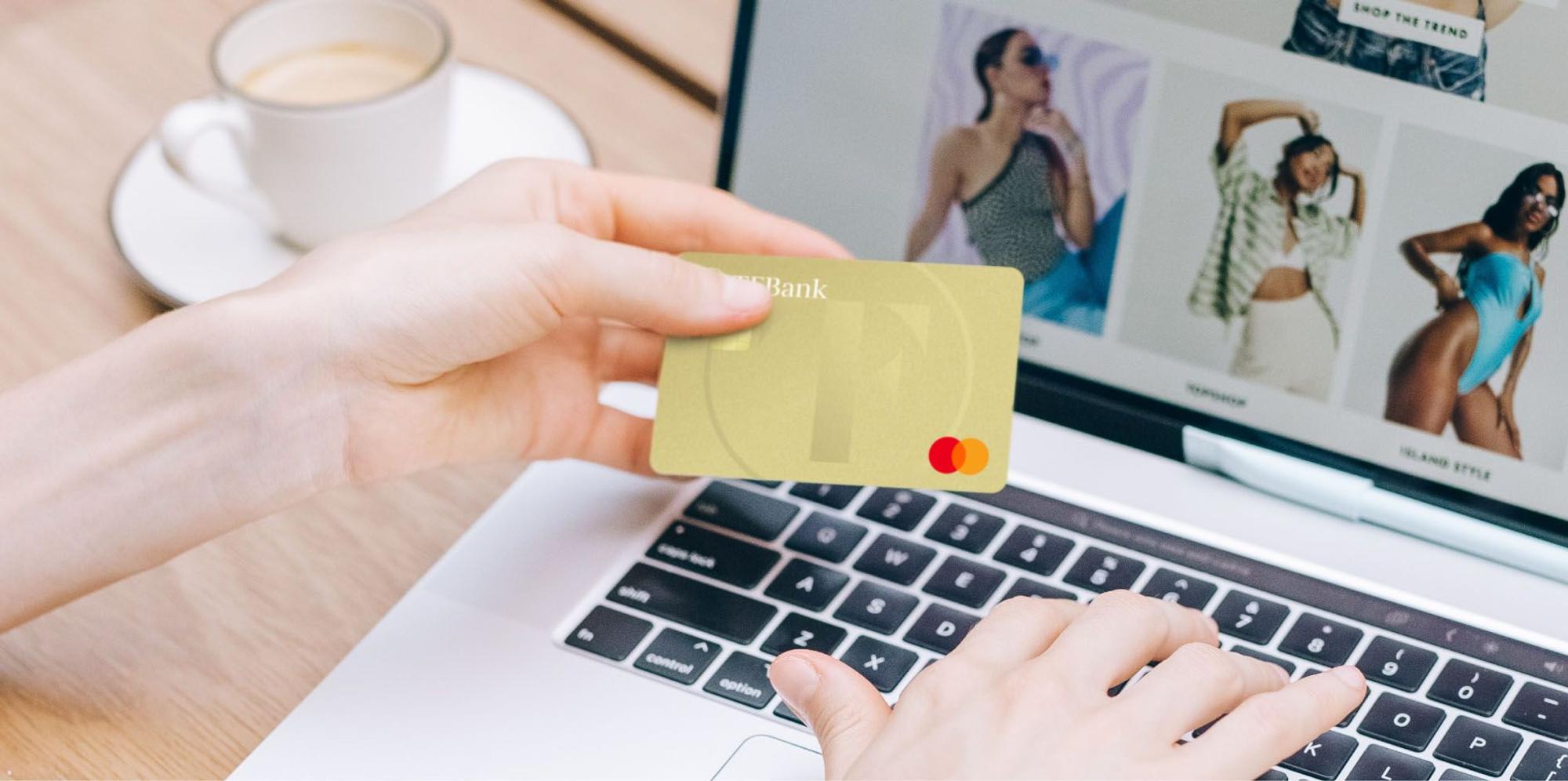 Eine weibliche Hand hält die Kreditkarte TF Mastercard Gold. Die andere Hand tippt etwas in den Laptop.
