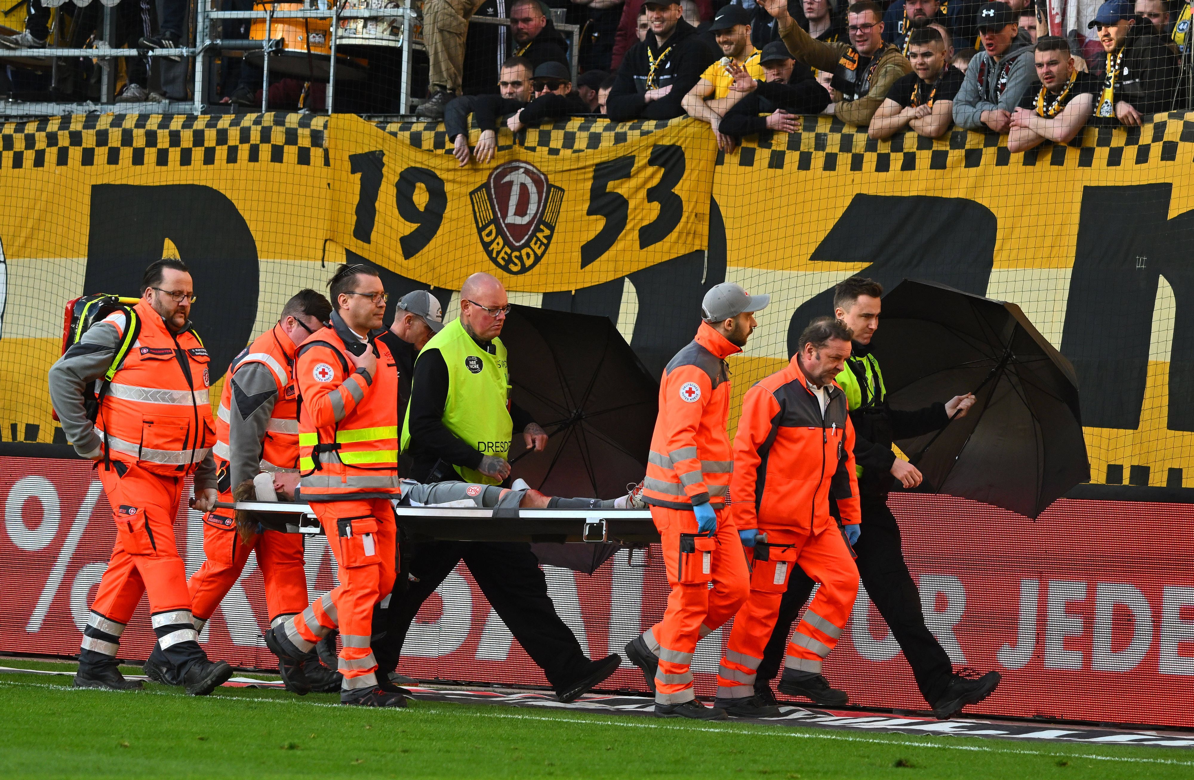 Felix Götze wird verletzt vom Platz getragen. Ordner schützen ihn mit Regenschirmen vor Wurfgeschossen.