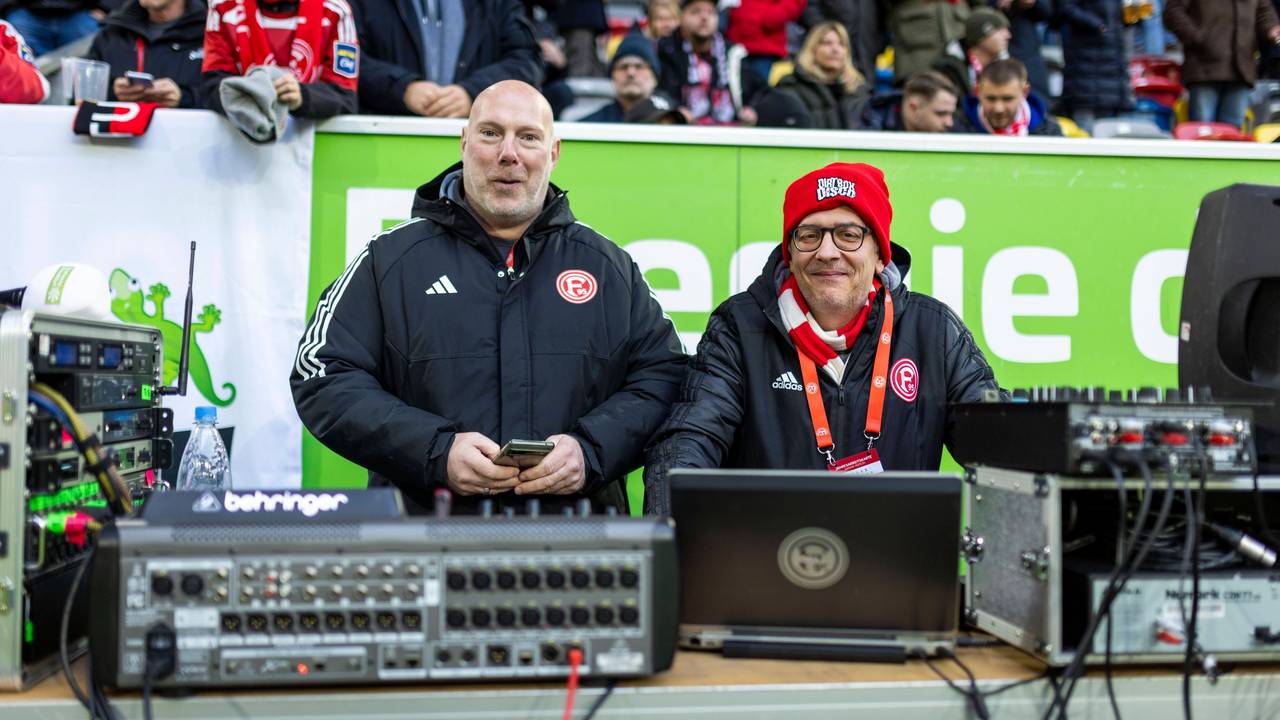 Fortuna Düsseldorfs Stadion-DJ Marcus „Opa“ Haefs und Techniker Carsten Johns lächeln beim Heimspiel gegen Holstein Kiel in die Kamera.