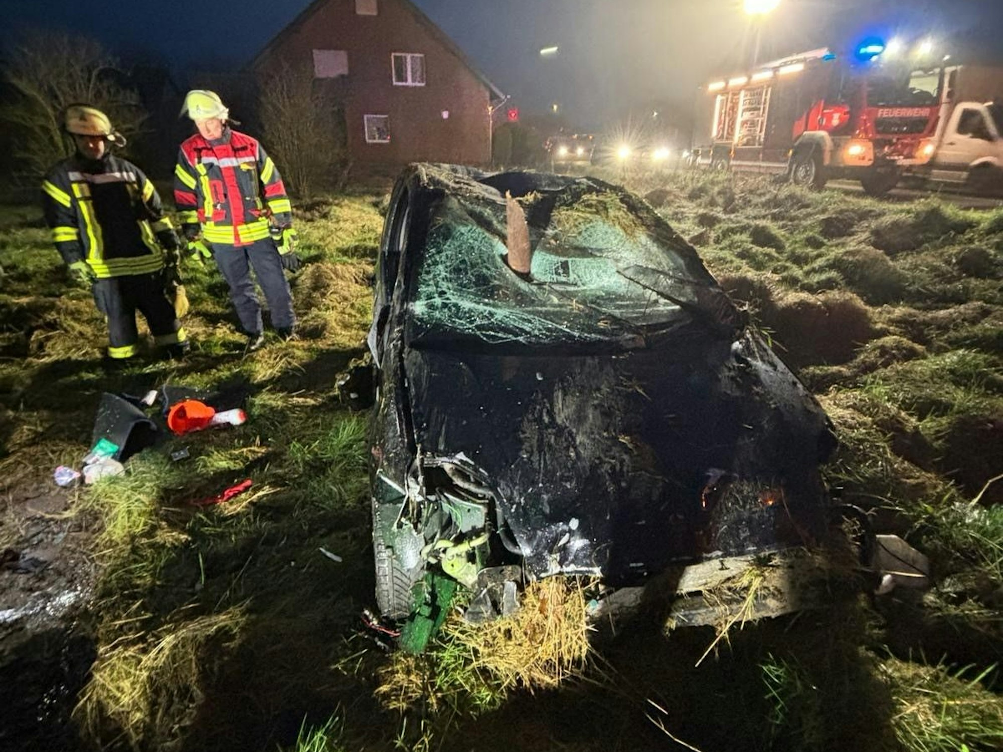 Ein Auto steht auf einer Wiese, aus seiner kaputten Windschutzscheibe ragt ein abgebrochener Holzpfosten. Neben dem Auto stehen zwei Feuerwehrmänner.