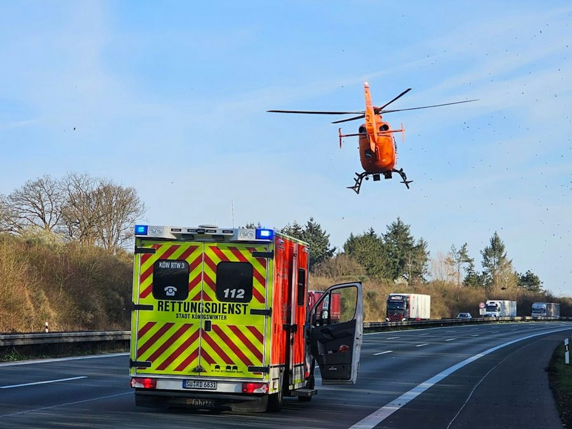 Ein Rettungshubschrauber ist über einer leeren Autobahn in der Luft, auf der Autobahn steht ein Rettungsfahrzeug.