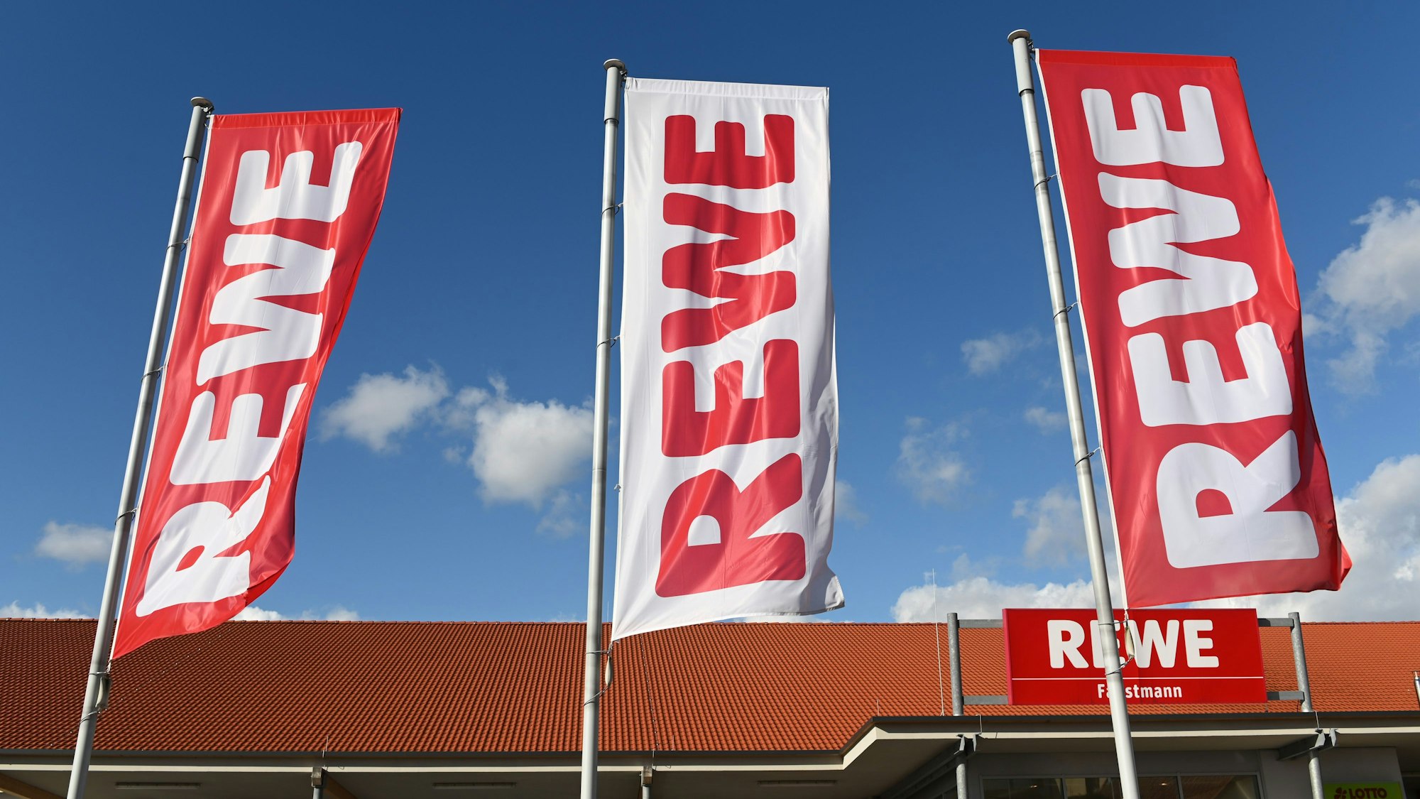 Fahnen mit dem Logo des Lebensmitteleinzelhändlers Rewe wehen vor einer Filiale.