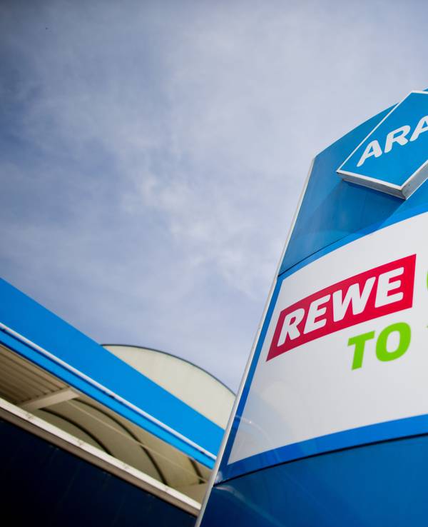 Die Logos von Rewe To Go und Aral stehen übereinander vor einer Tankstelle.