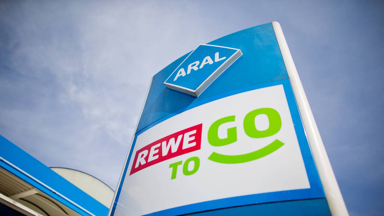 Die Logos von Rewe To Go und Aral stehen übereinander vor einer Tankstelle.