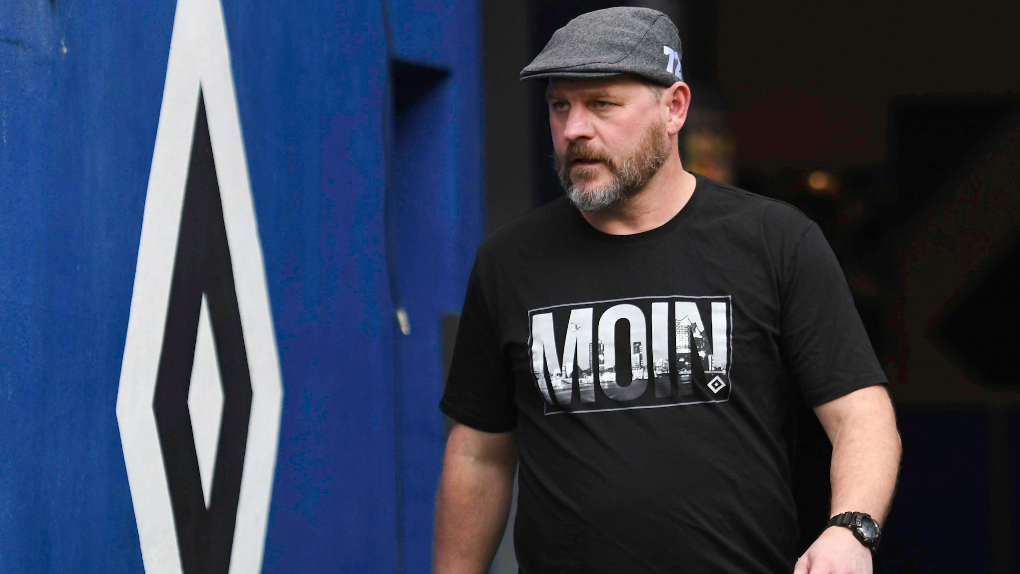 Steffen Baumgart geht mit Schiebermütze und T-Shirt aus der Kabine auf den Platz.