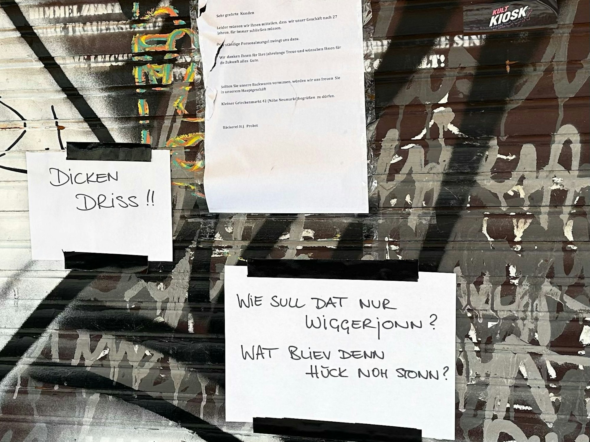 Zettel mit den Aufschriften „Dicken Driss!“ und „Wie sull dat nur wigger jonn? Wat bliev denn hück noh stonn?“ hängen an der geschlossenen Bäckerei Probst.