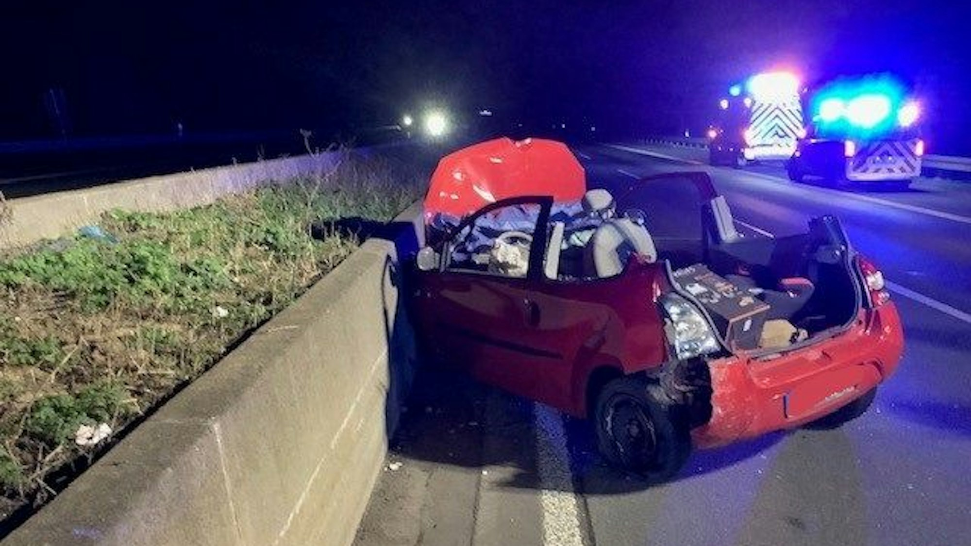 Ein stark beschädigtes Auto ohne Dach steht auf der Autobahn an einer niedrigen Betonwand.