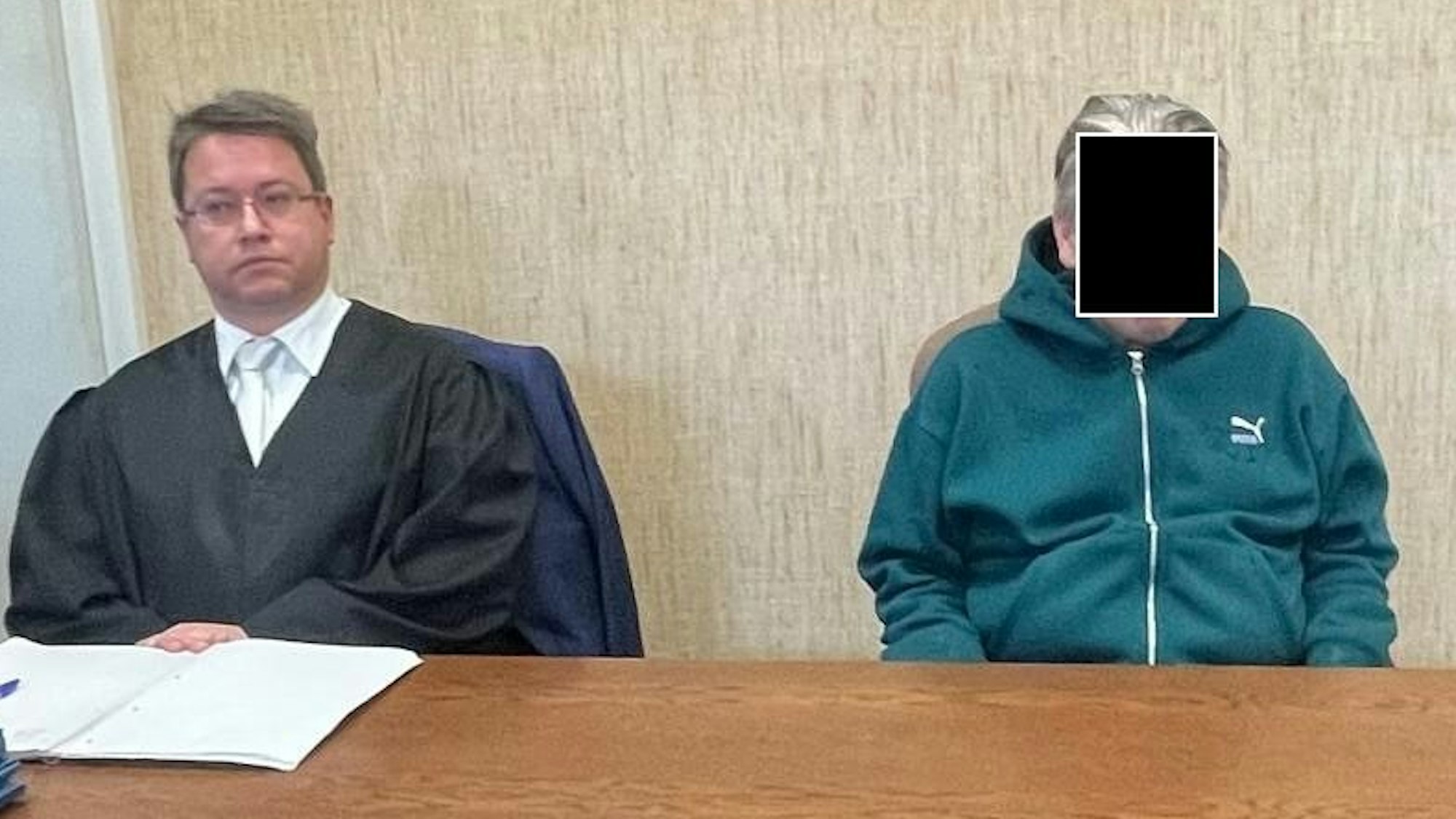 Ein Mann in einer Trainingsjacke sitzt an einem Tisch neben einem Anwalt.