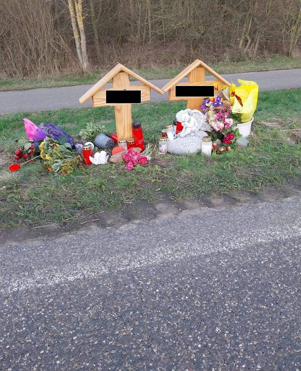 An zwei Holzkreuzen liegen Blumen, brennen Kerzen.