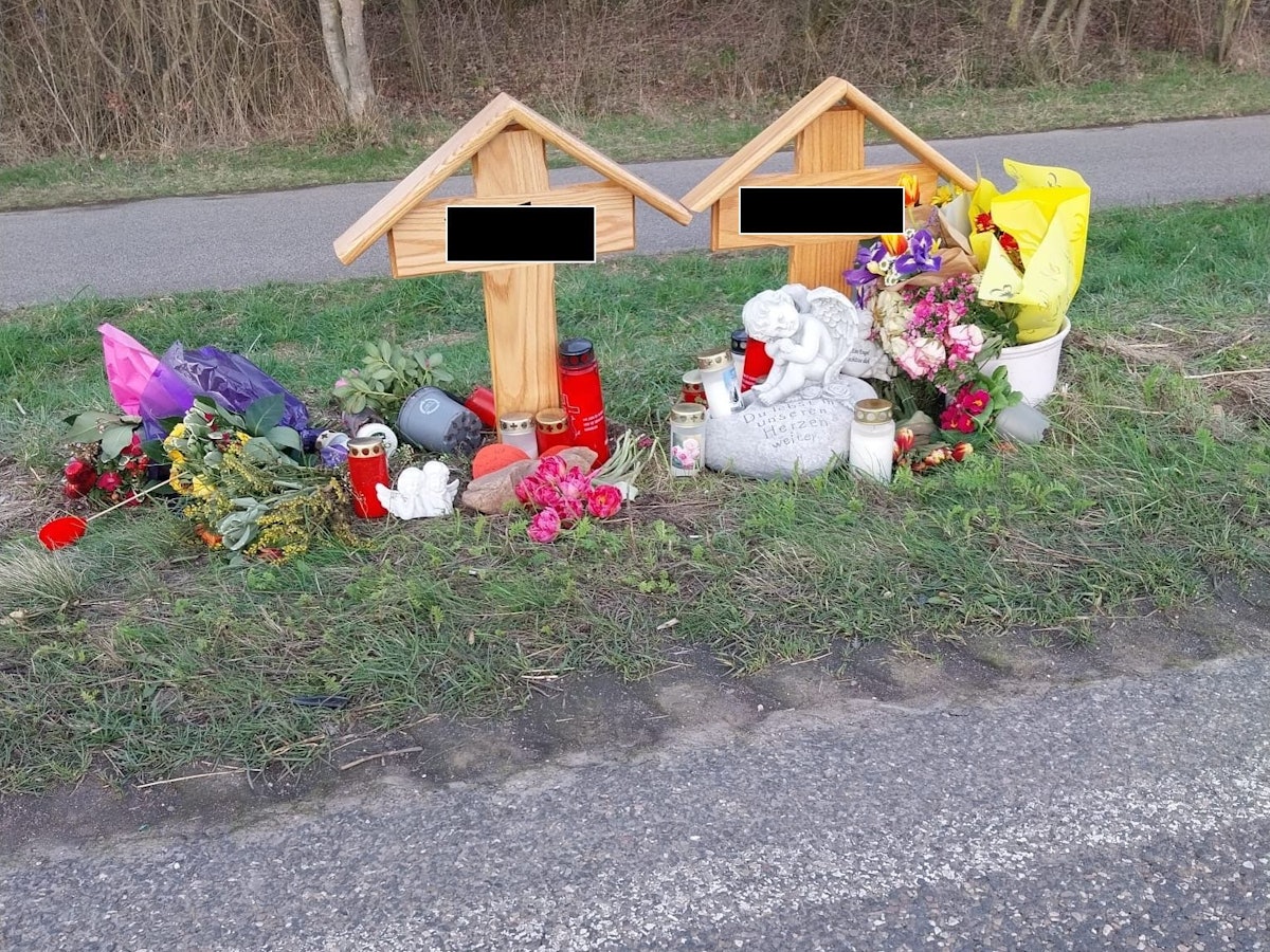 An zwei Holzkreuzen liegen Blumen, brennen Kerzen.