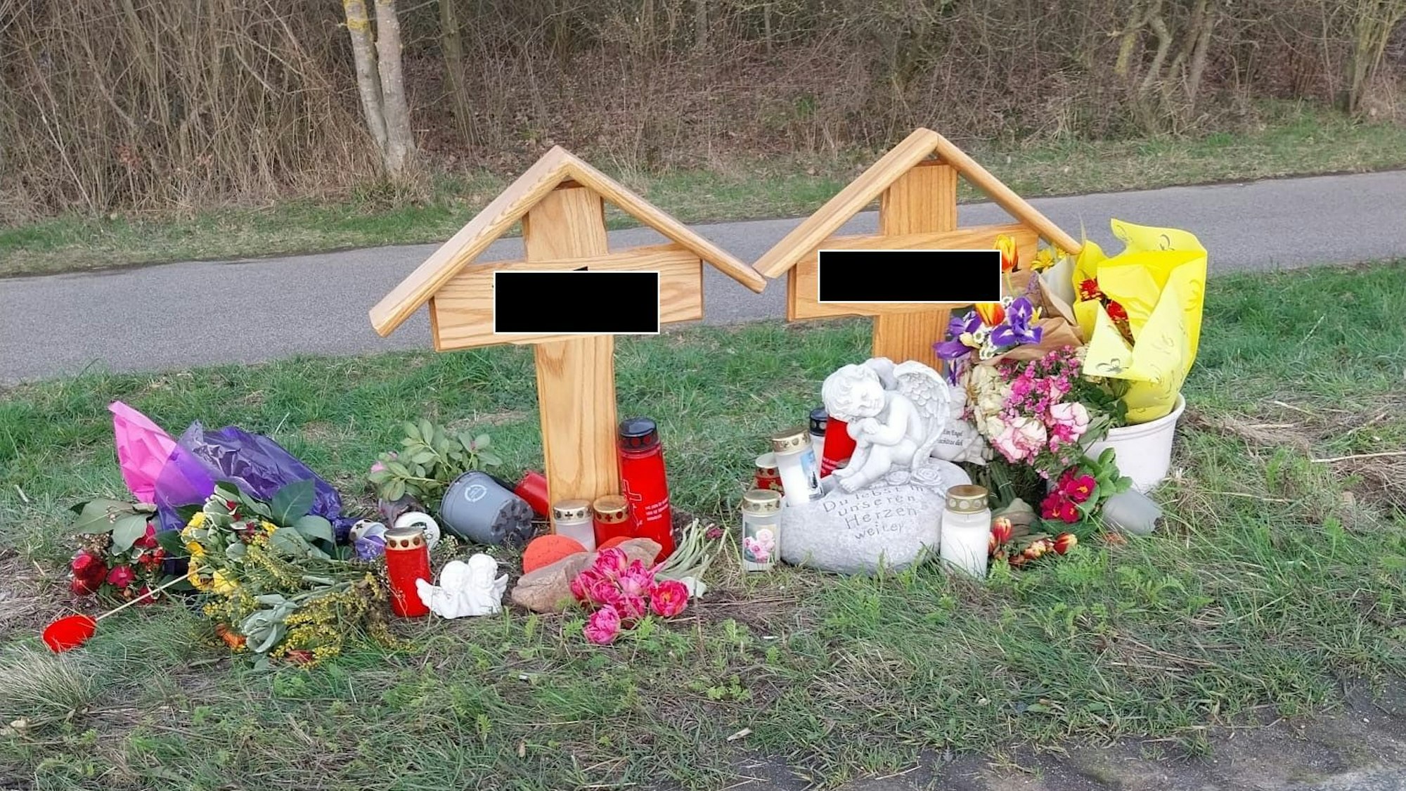 An zwei Holzkreuzen liegen Blumen, brennen Kerzen.