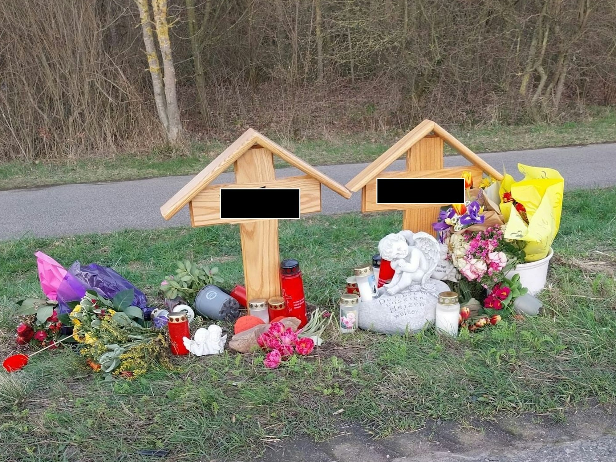 Zwei Holzkreuze stehen am Fahrbahnrand, davor Blumen, Kerzen und ein Engel aus Stein.