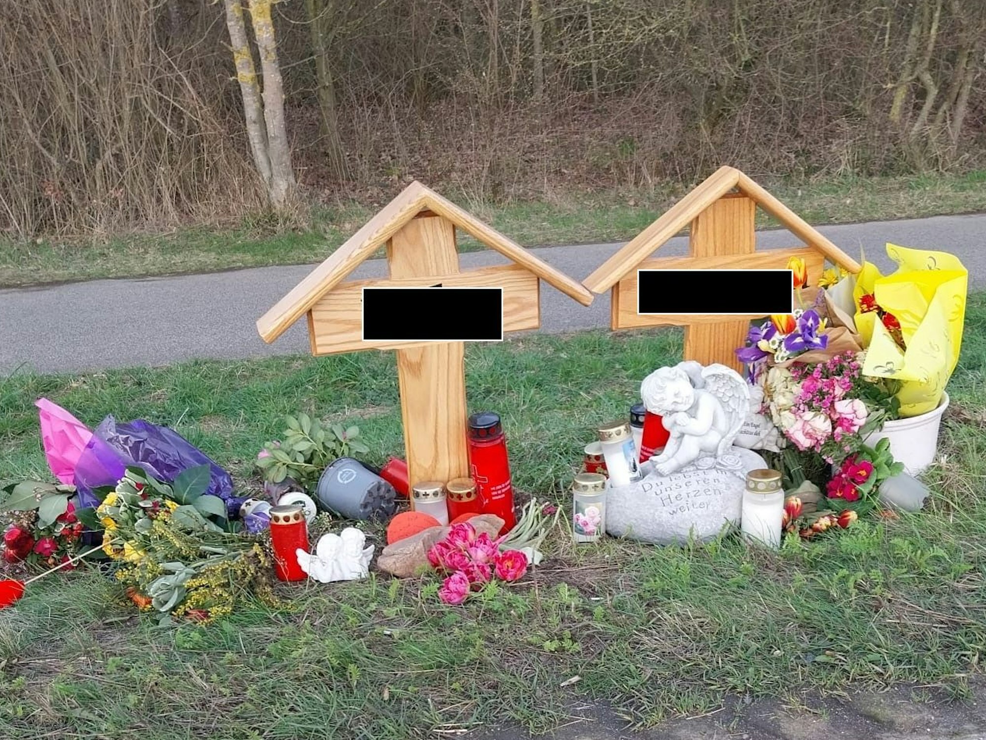 An einer Straße stehen zwei Holzkreuze, daneben Blumen, Kerzen und Engelsfiguren.