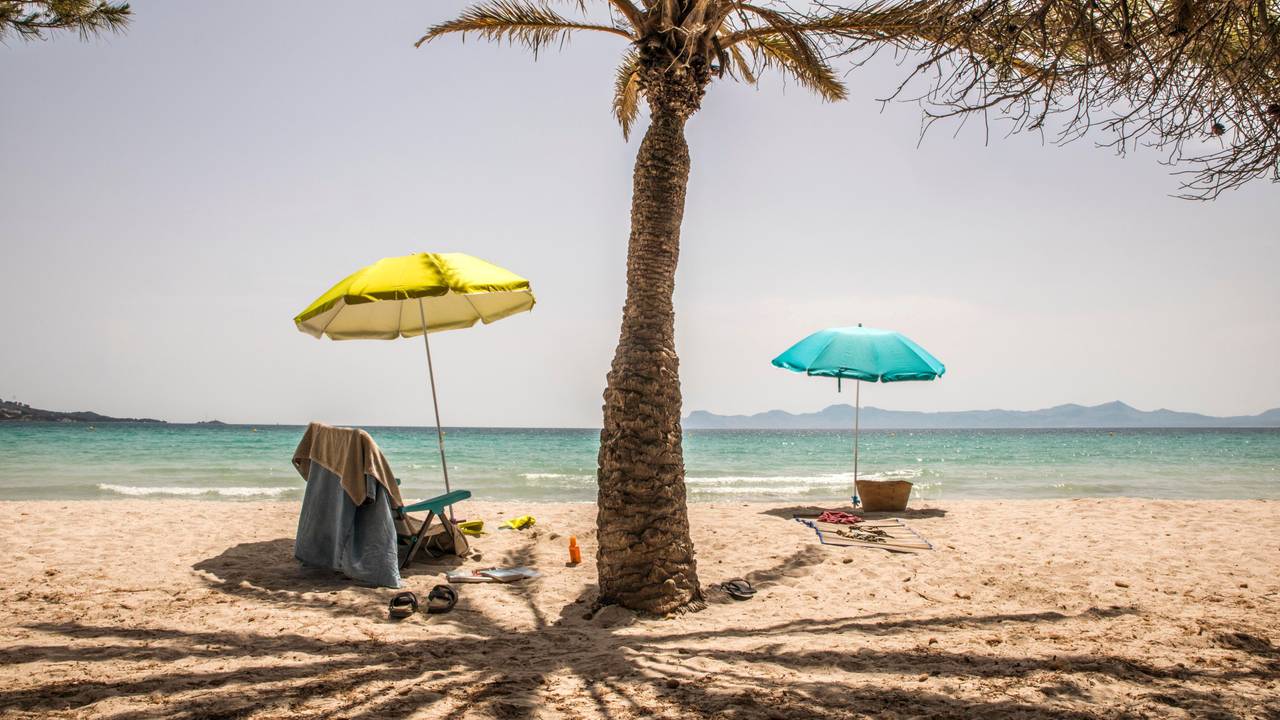 Zwei leere Stühle unter Sonnenschirmen stehen bei Sonnenschein an einem Strand.