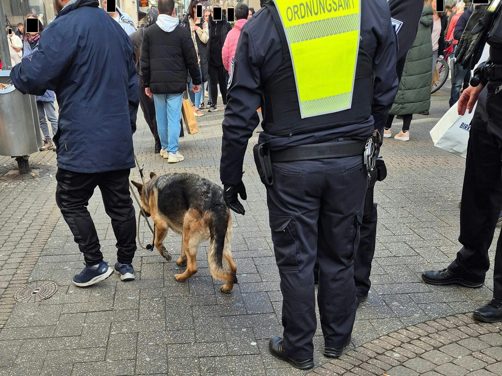 Ein Mitarbeiter des Ordnungsamtes steht auf der belebten Schildergasse, vor ihm führt ein Mann einen Schäferhund an der Leine.