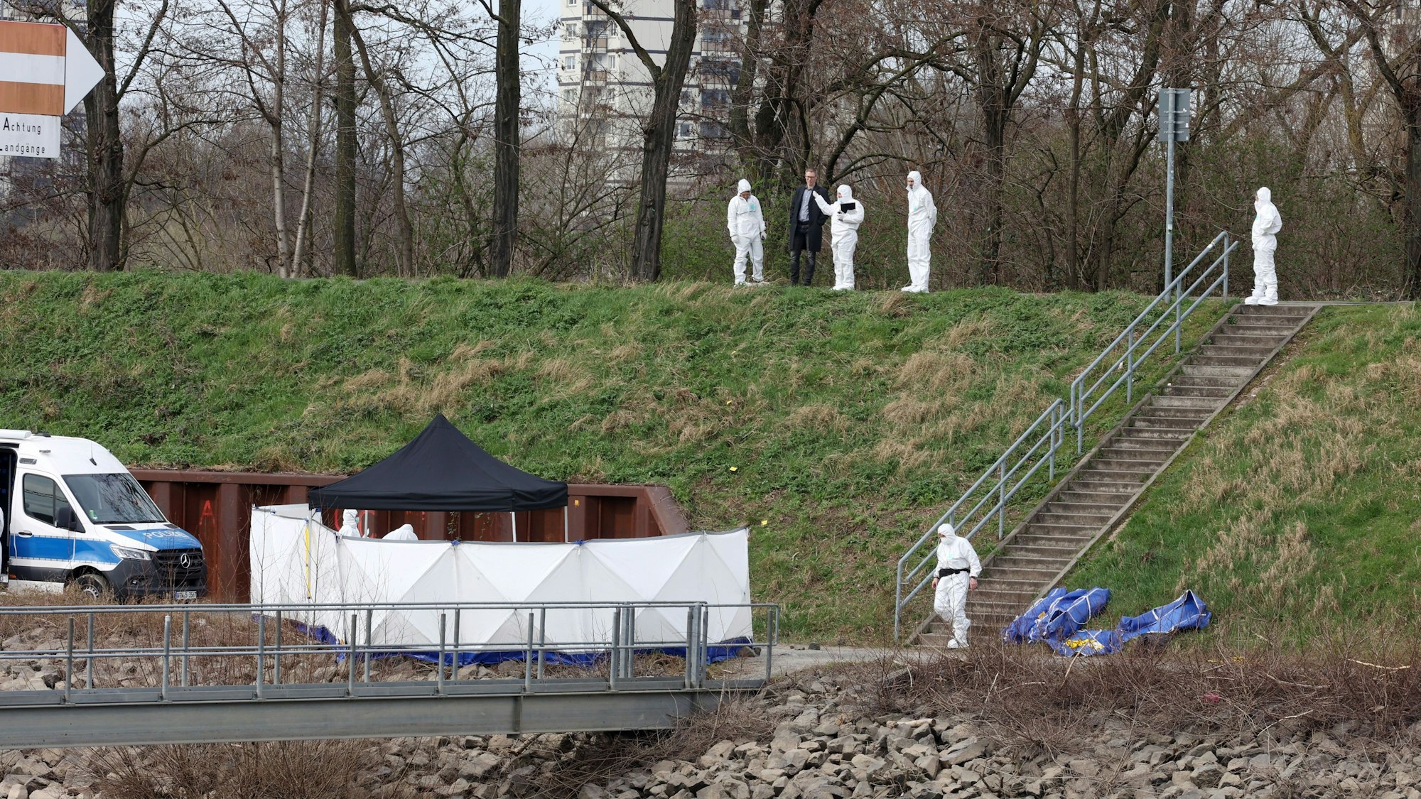 Am Fuß eines Abhangs steht ein Zelt und ein Sichtschutz, dem sich ein Mensch in einem Schutzanzug nähert. Vier weitere in Schutzanzügen sowie ein Mann in einem Anzug stehen oberhalb.