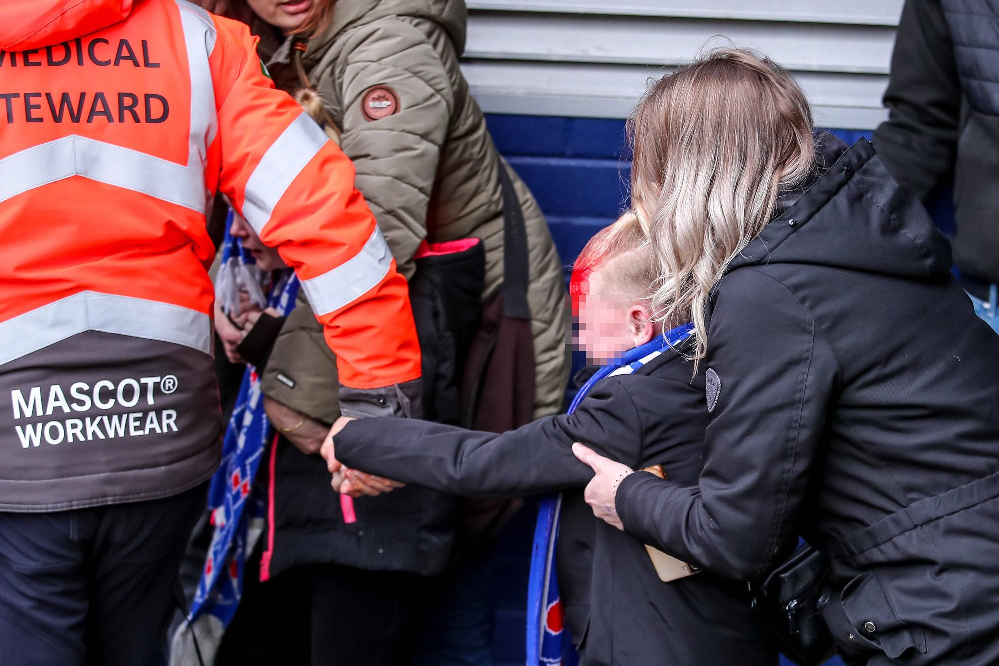 Ein am Kopf verletztes Kind wird von einem Ordner an der Hand aus dem Stadion geführt. Von hinten hält eine Frau das Kind fest.