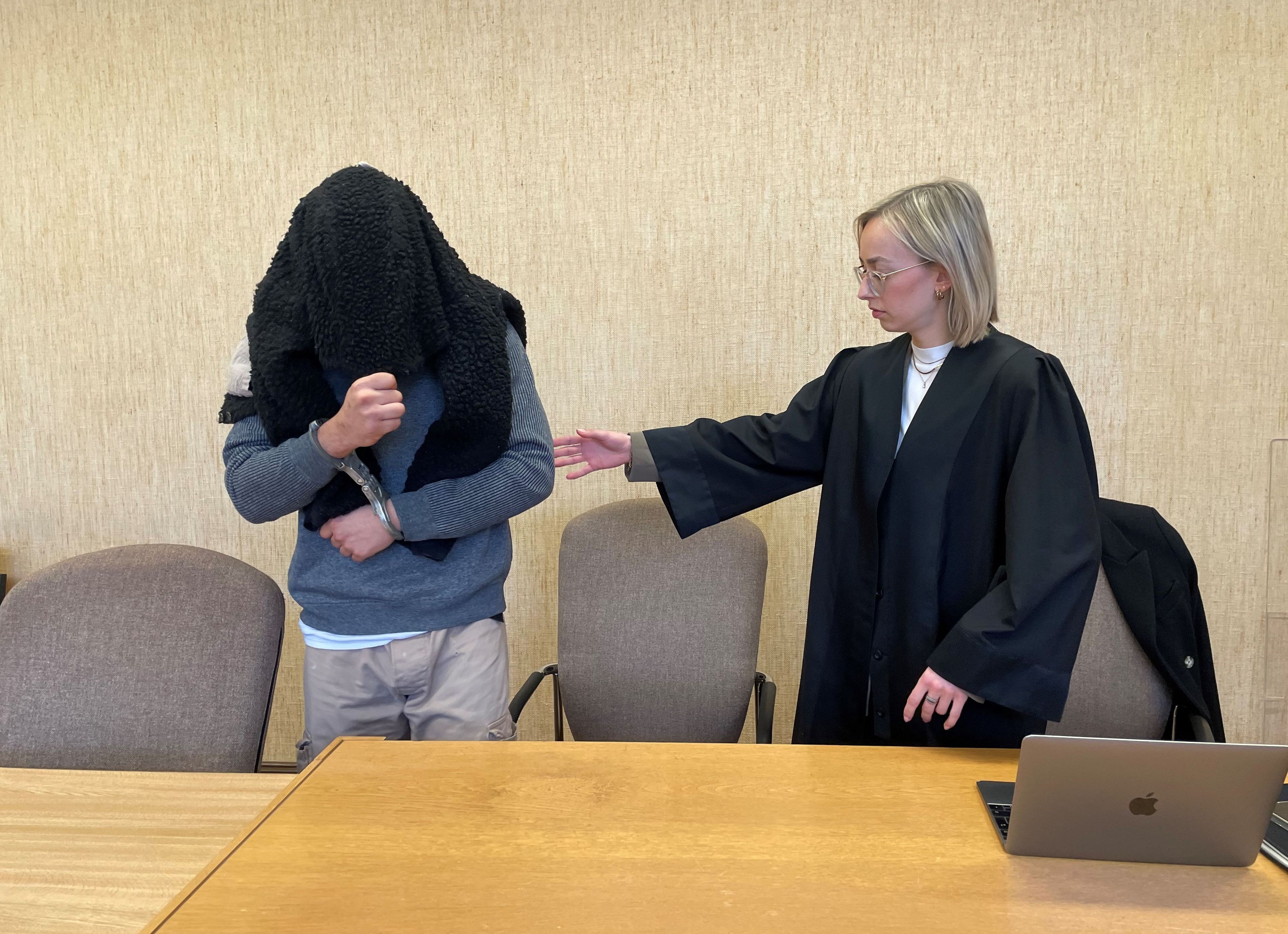 Ein Mann in Handschellen und mit einem Pullover über Kopf und Gesicht nimmt auf der Anklagebank Platz. Seine Anwältin leitet ihn mit der Hand.