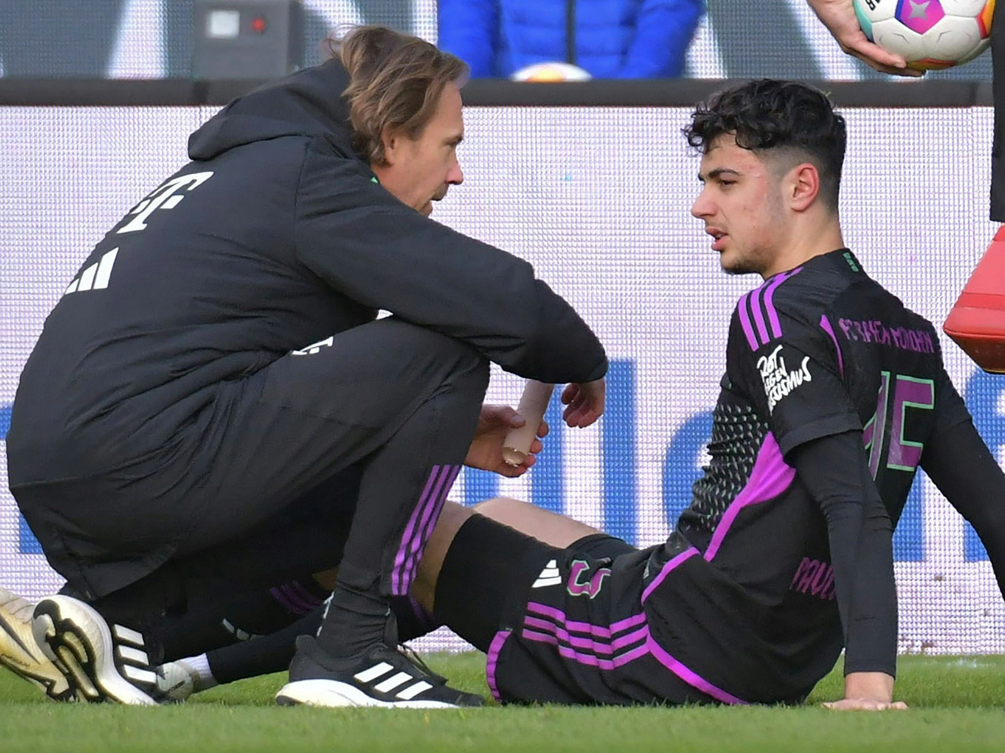 Aleksandar Pavlovic sitzt bei der Bundesliga-Partie in Darmstadt mit geschwollenem Auge auf dem Boden und wird von Arzt Dr. Peter Ueblacker behandelt.