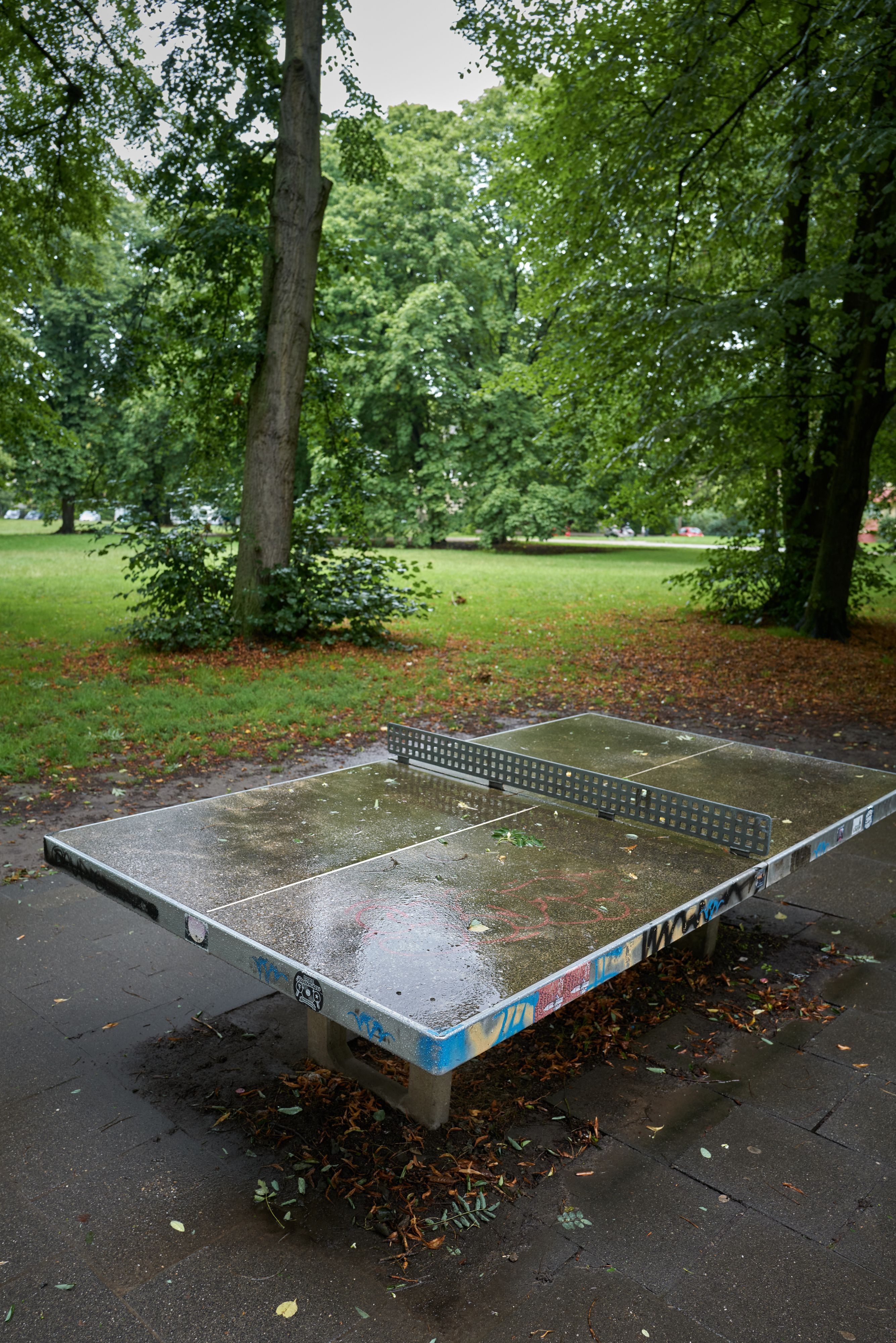 Eine Tischtennisplatte steht auf einer asphaltierten Fläche in einem Park.