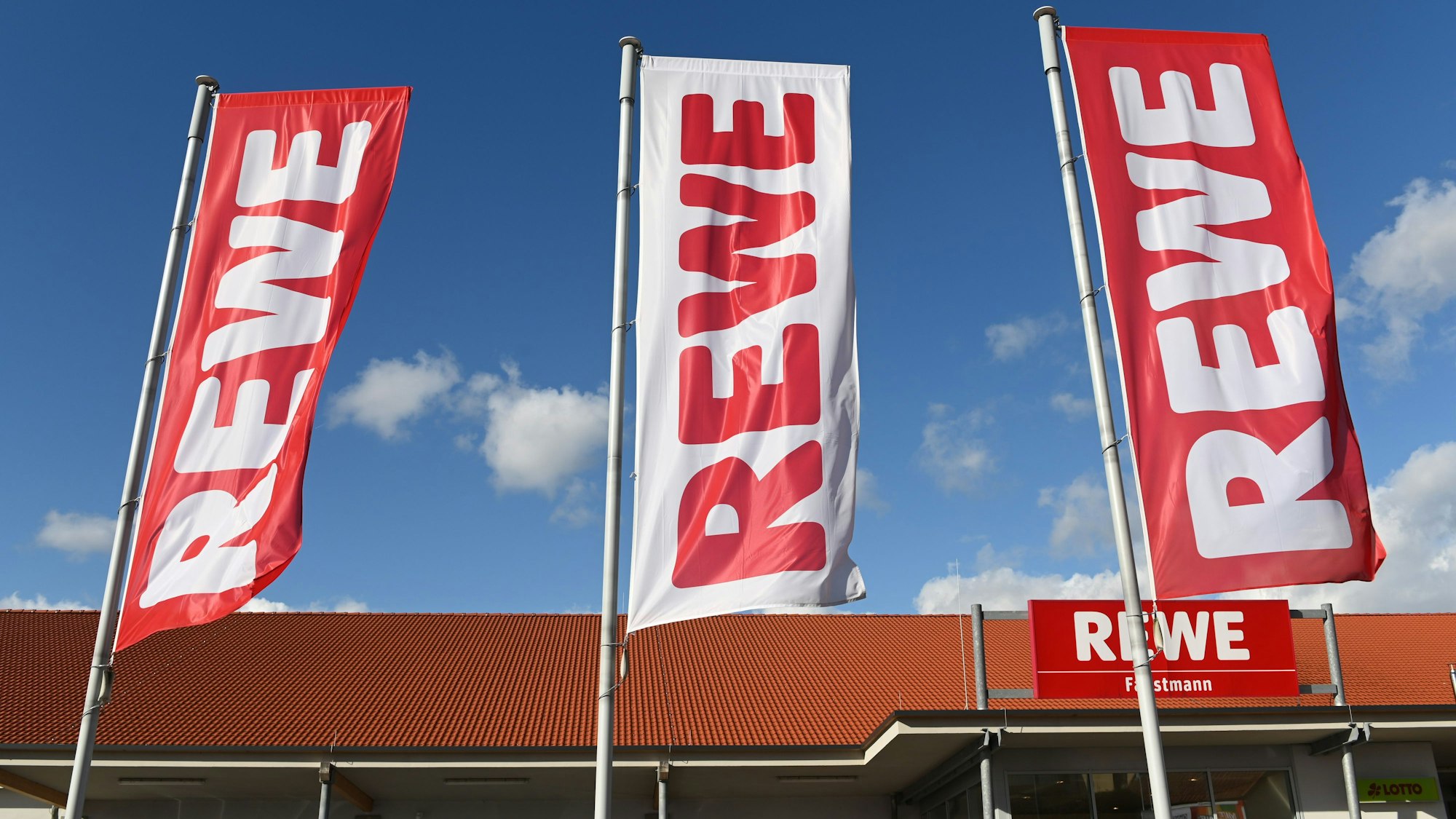Fahnen mit dem Rewe Logo wehen vor einer Filiale.