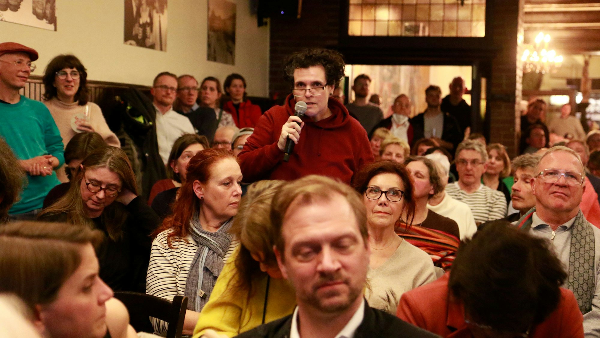 Menschen sitzen und stehen gedrängt in einer Gaststätte, ein Mann hält ein Mikrofon.