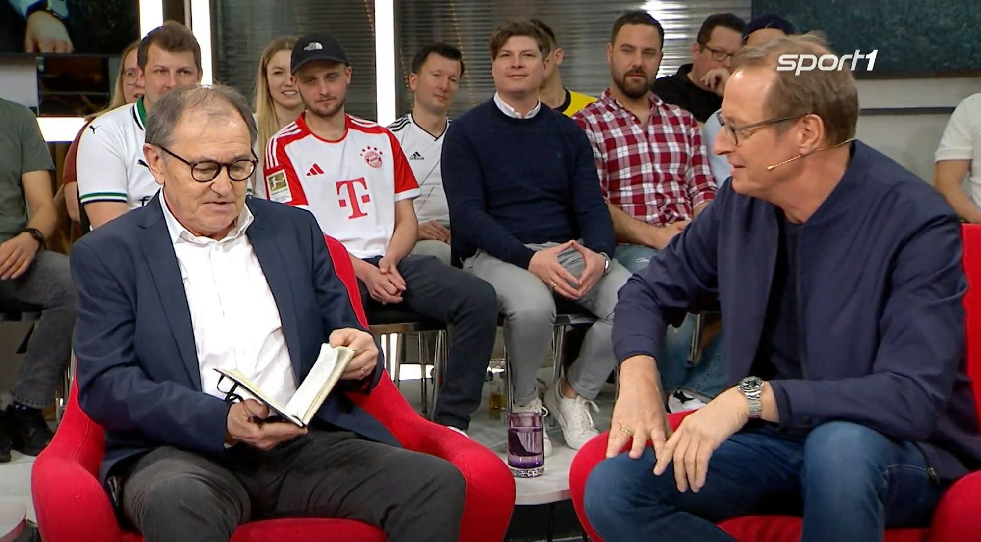Ewald Lienen schaut beim Sport1-„Doppelpass“ in sein Notizbuch, auf das ihn Florian König gerade angesprochen hat.