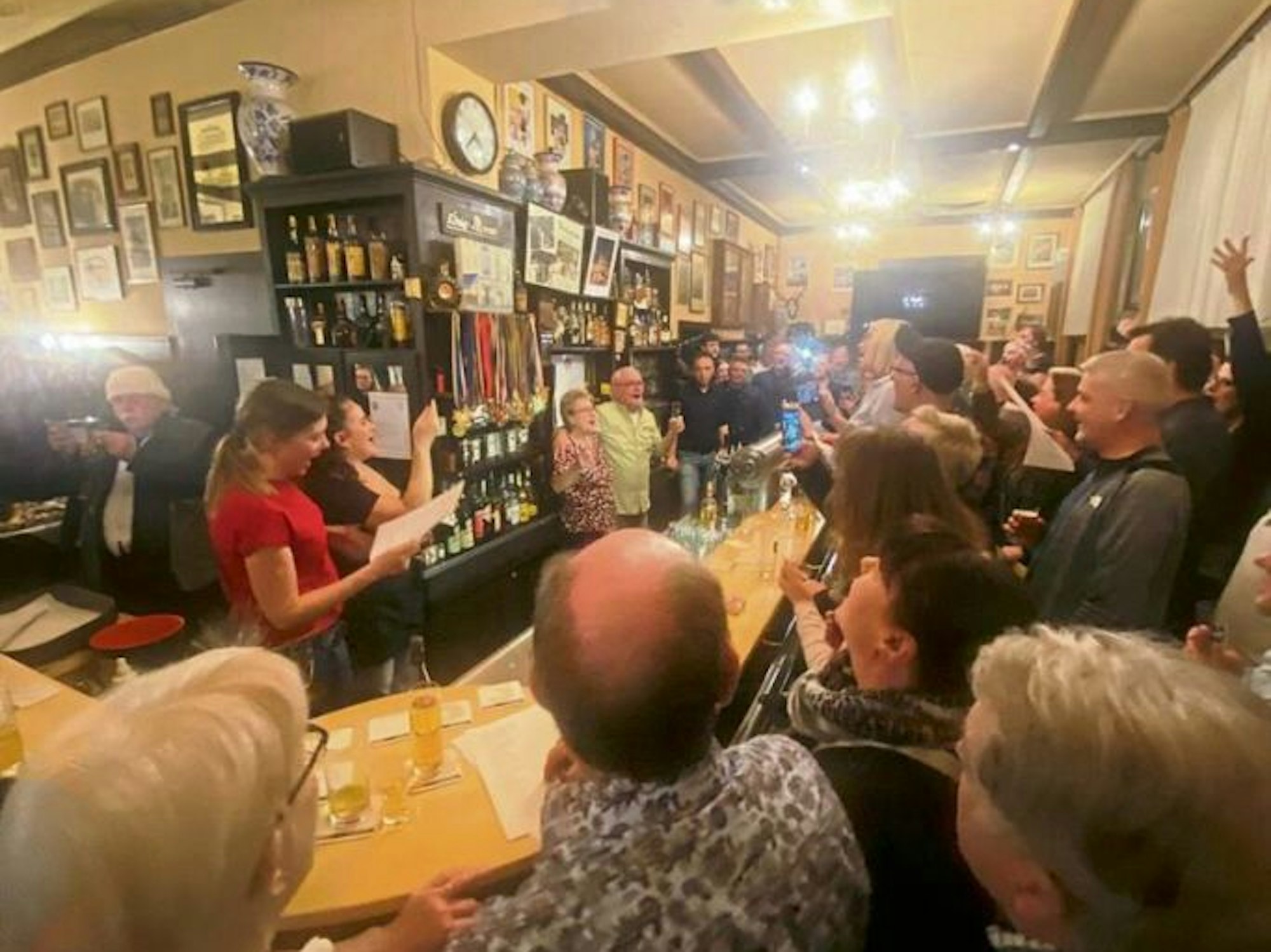 Das Wirtsehepaar steht hinter der Theke, vor der Theke erheben zahlreiche Gäste ihre Gläser.