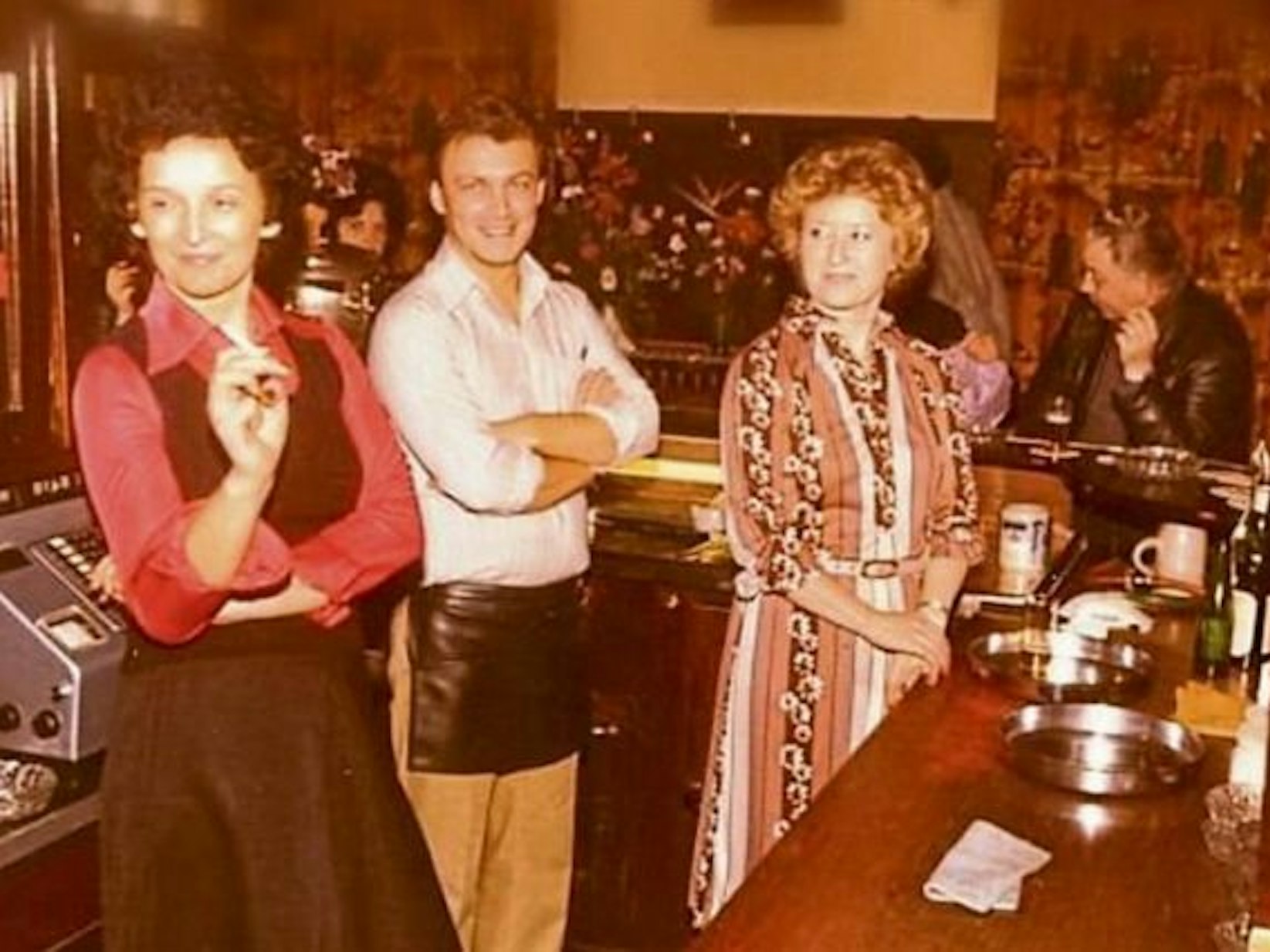 Auf einem uralten Foto stehen zwei Frauen und ein Mann hinter einer Kneipen-Theke, eine der Frauen raucht.
