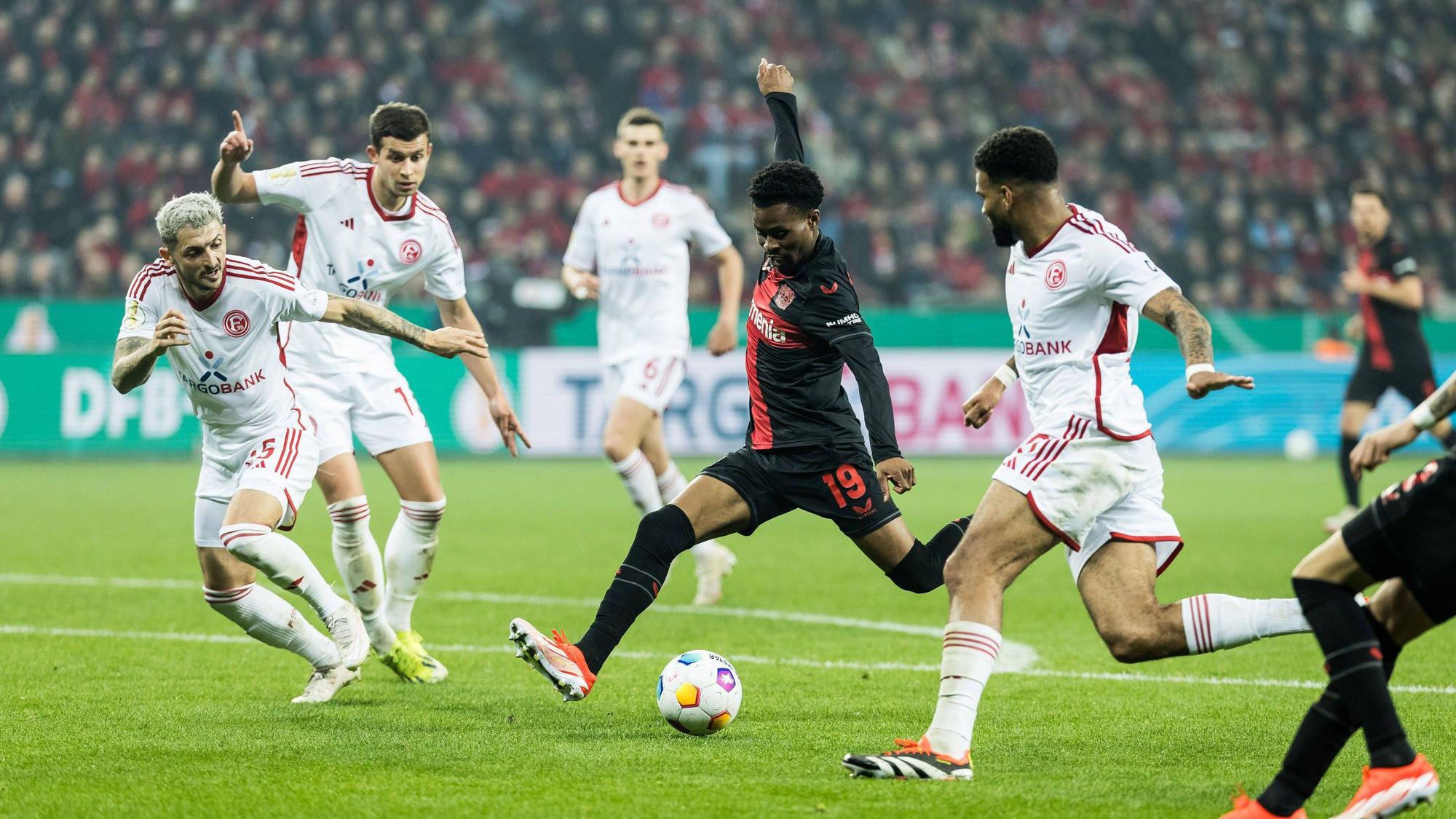 Nathan Tella schießt den Ball. Fortuna Düsseldorf Spieler stehen drumherum: Von l. n. r.: Matthias Zimmermann, Tim Oberdorf, Jamil Siebert.