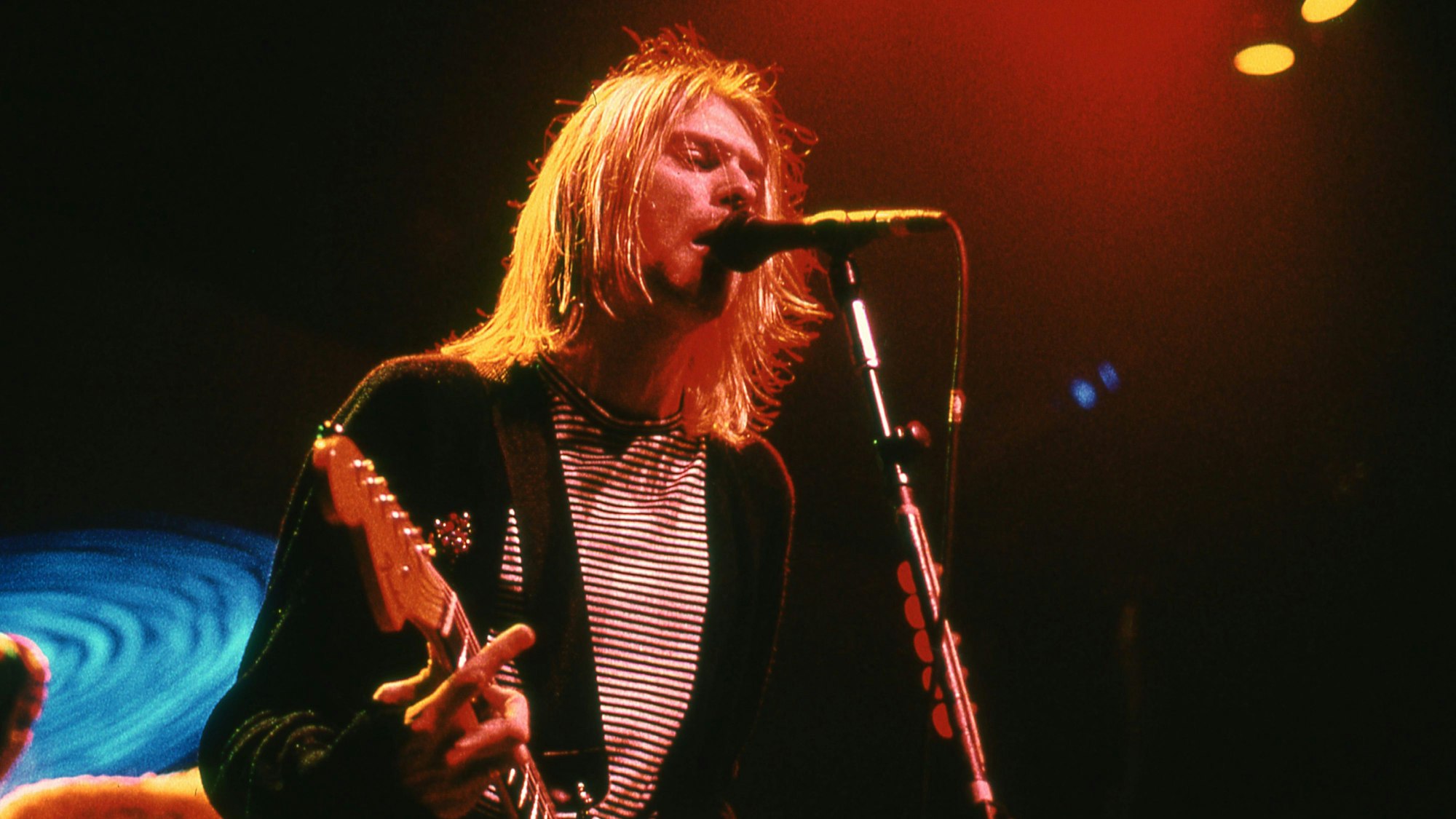 Kurt Cobain steht mit langen Haaren und seiner Gitarre vor einem Mikrofon und singt.