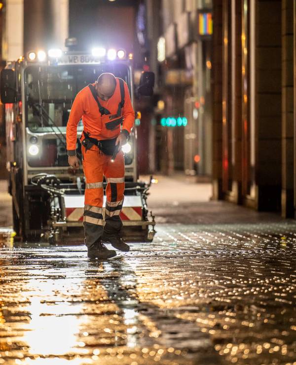 Ein Mitarbeiter der AWB checkt nachts den Boden in der Kölner Innenstadt, hinter ihm fährt eine AWB-Maschine.