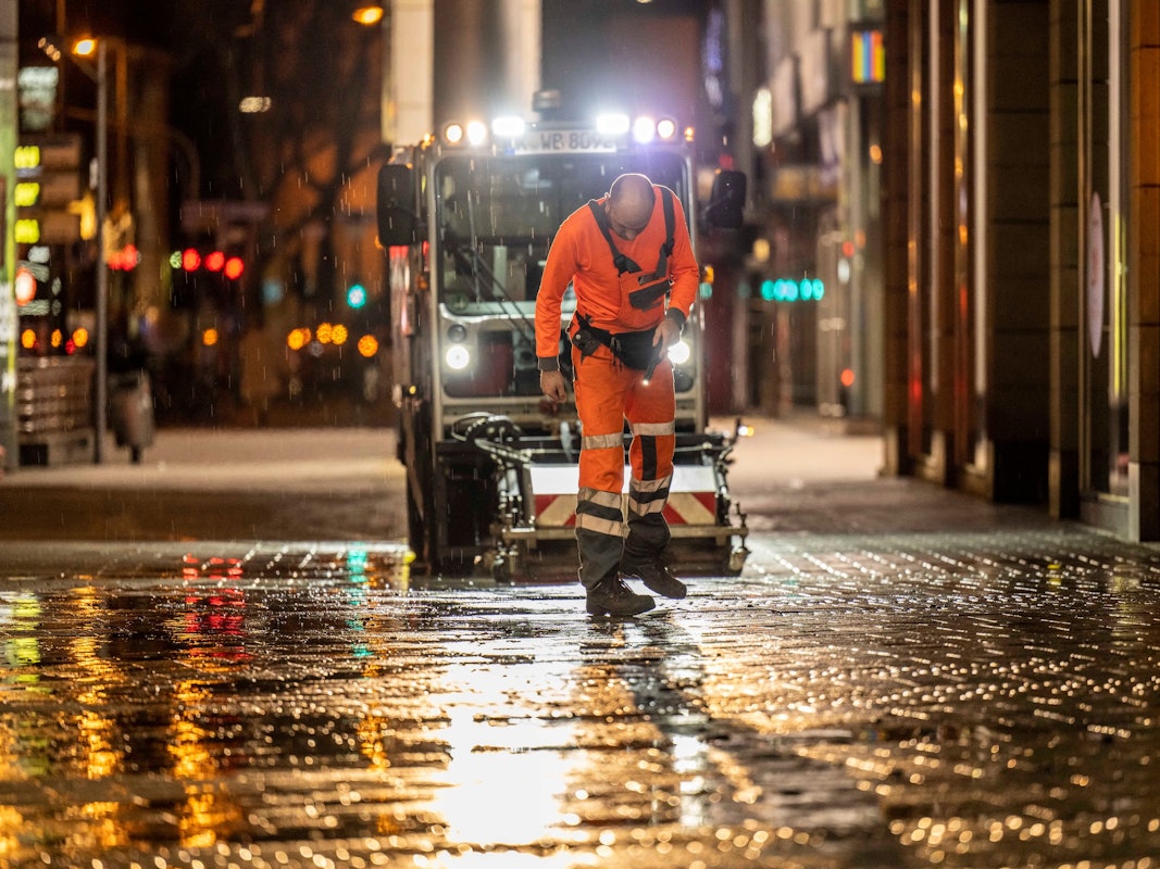 Ein Mitarbeiter der AWB checkt nachts den Boden in der Kölner Innenstadt, hinter ihm fährt eine AWB-Maschine.