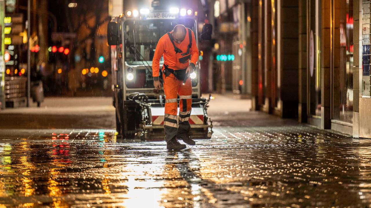 Ein Mitarbeiter der AWB checkt nachts den Boden in der Kölner Innenstadt, hinter ihm fährt eine AWB-Maschine.