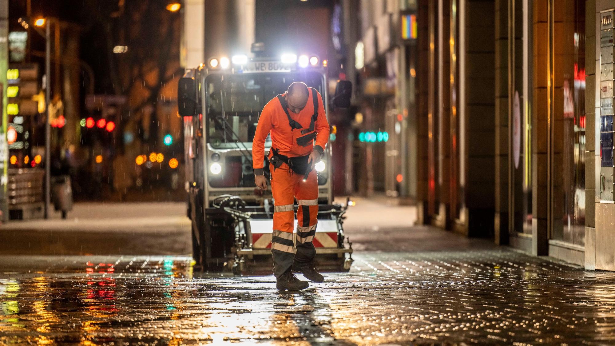Ein Mitarbeiter der AWB checkt nachts den Boden in der Kölner Innenstadt, hinter ihm fährt eine AWB-Maschine.