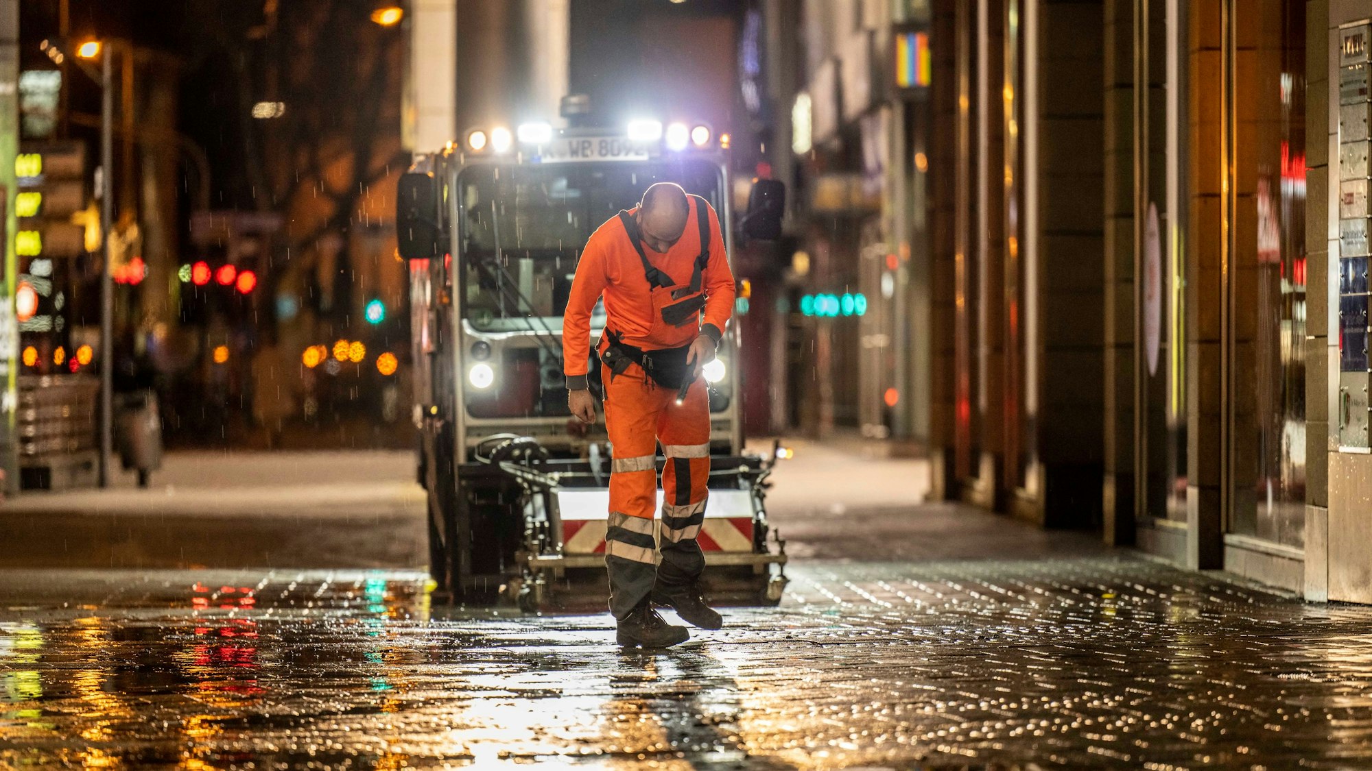 Ein Mitarbeiter der AWB checkt nachts den Boden in der Kölner Innenstadt, hinter ihm fährt eine AWB-Maschine.