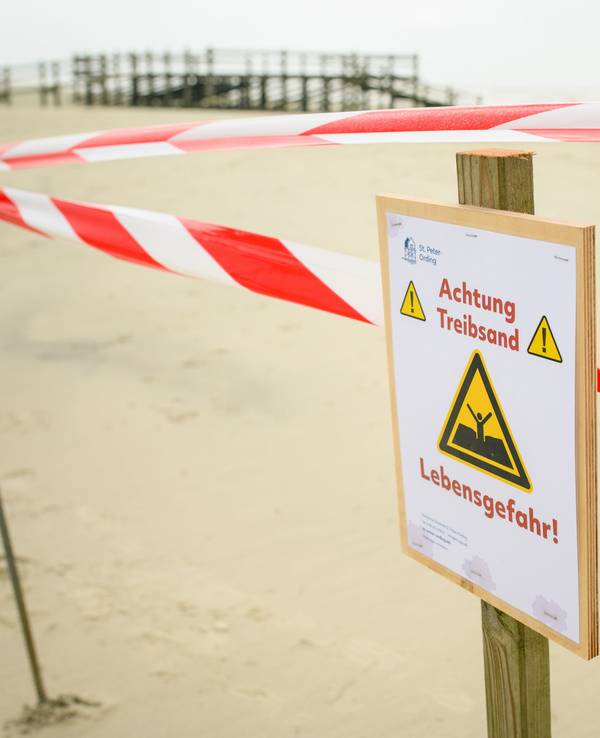 Flatterband und ein Warnhinweis („Achtung Treibsand – Lebensgefahr!“) sind an einem abgesperrten Bereich auf dem Strand von St. Peter-Ording zu sehen.