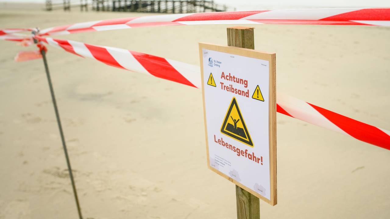 Flatterband und ein Warnhinweis („Achtung Treibsand – Lebensgefahr!“) sind an einem abgesperrten Bereich auf dem Strand von St. Peter-Ording zu sehen.