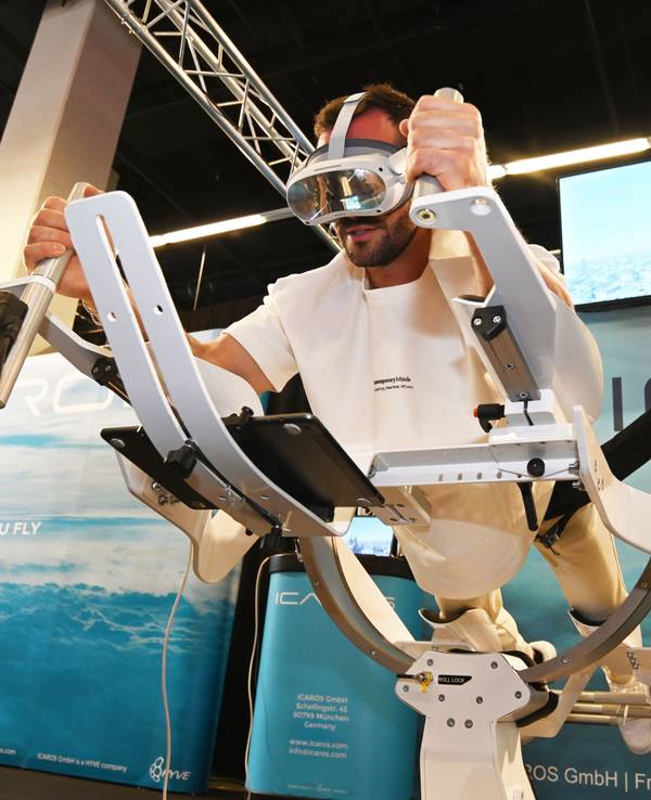 Personal Trainer Manuel Gajus liegt in Supermann-Pose auf dem ICAROS Gerät und hat eine VR-Brille an. Auf dem Bildschirm im Hintergrund sieht man den Kölner Dom.
