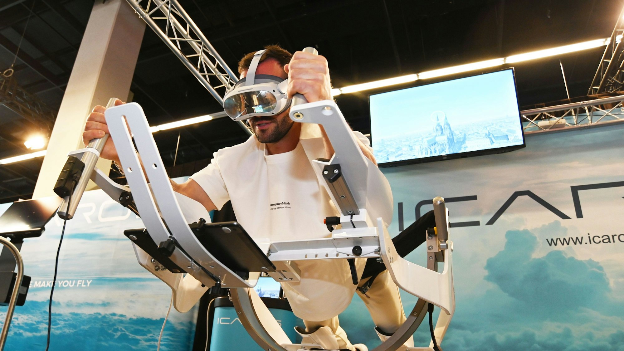 Personal Trainer Manuel Gajus liegt in Supermann-Pose auf dem ICAROS Gerät und hat eine VR-Brille an. Auf dem Bildschirm im Hintergrund sieht man den Kölner Dom.