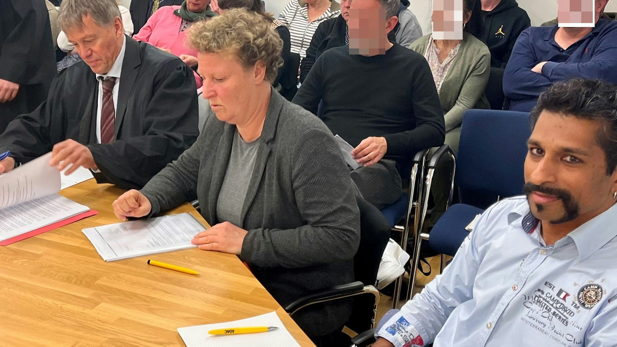 Eine Frau sitzt zwischen zwei Männern an einem Tisch, im Hintergrund Publikum.