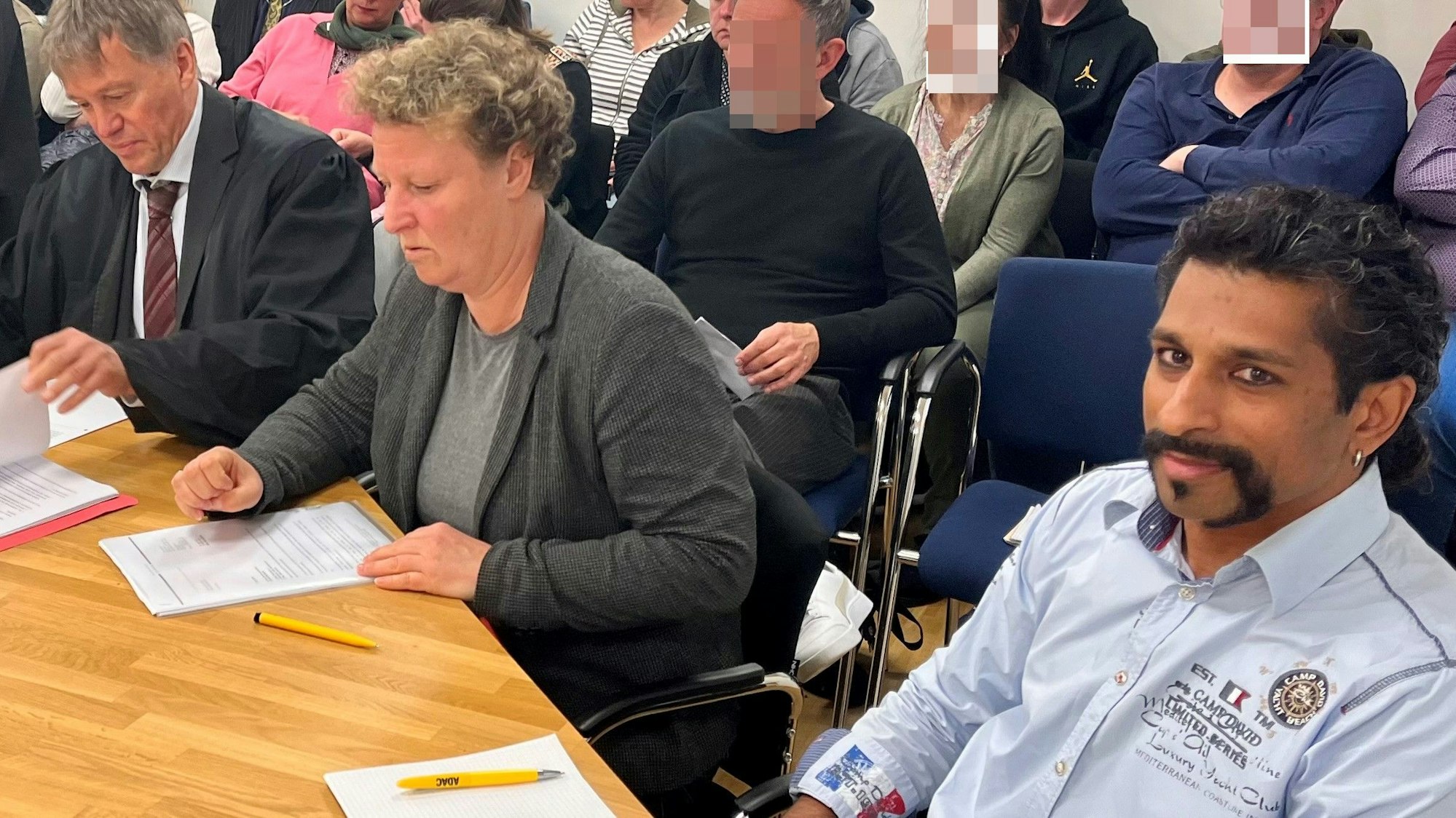 Eine Frau sitzt zwischen zwei Männern an einem Tisch, hinter ihnen mehrere vollbesetzte Stuhlreihen.