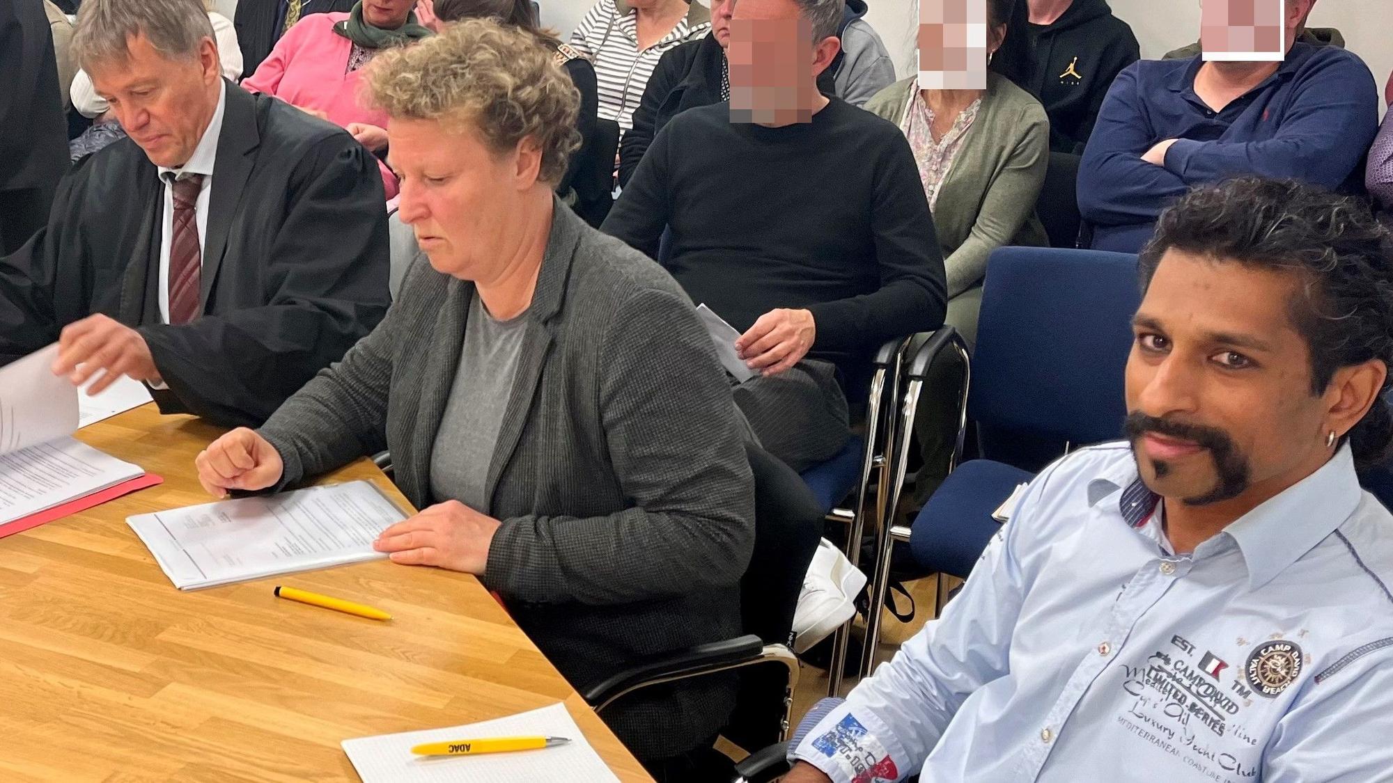 Zwei Männer und eine Frau sitzen an einem Tisch, im Hintergrund weitere Personen.
