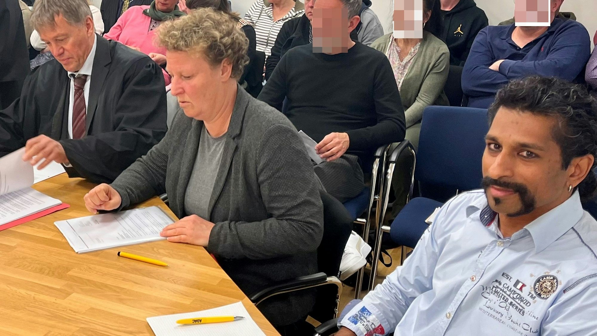 Zwei Männer und eine Frau sitzen an einem Tisch, im Hintergrund weitere Personen.