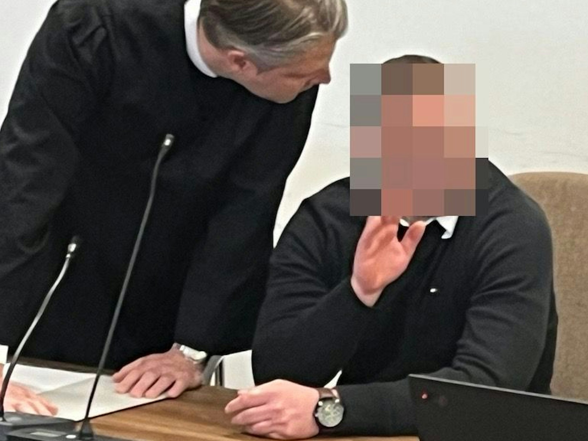 Ein Mann sitzt auf der Anklagebank und winkt, sein Anwalt beugt sich zu ihm runter.