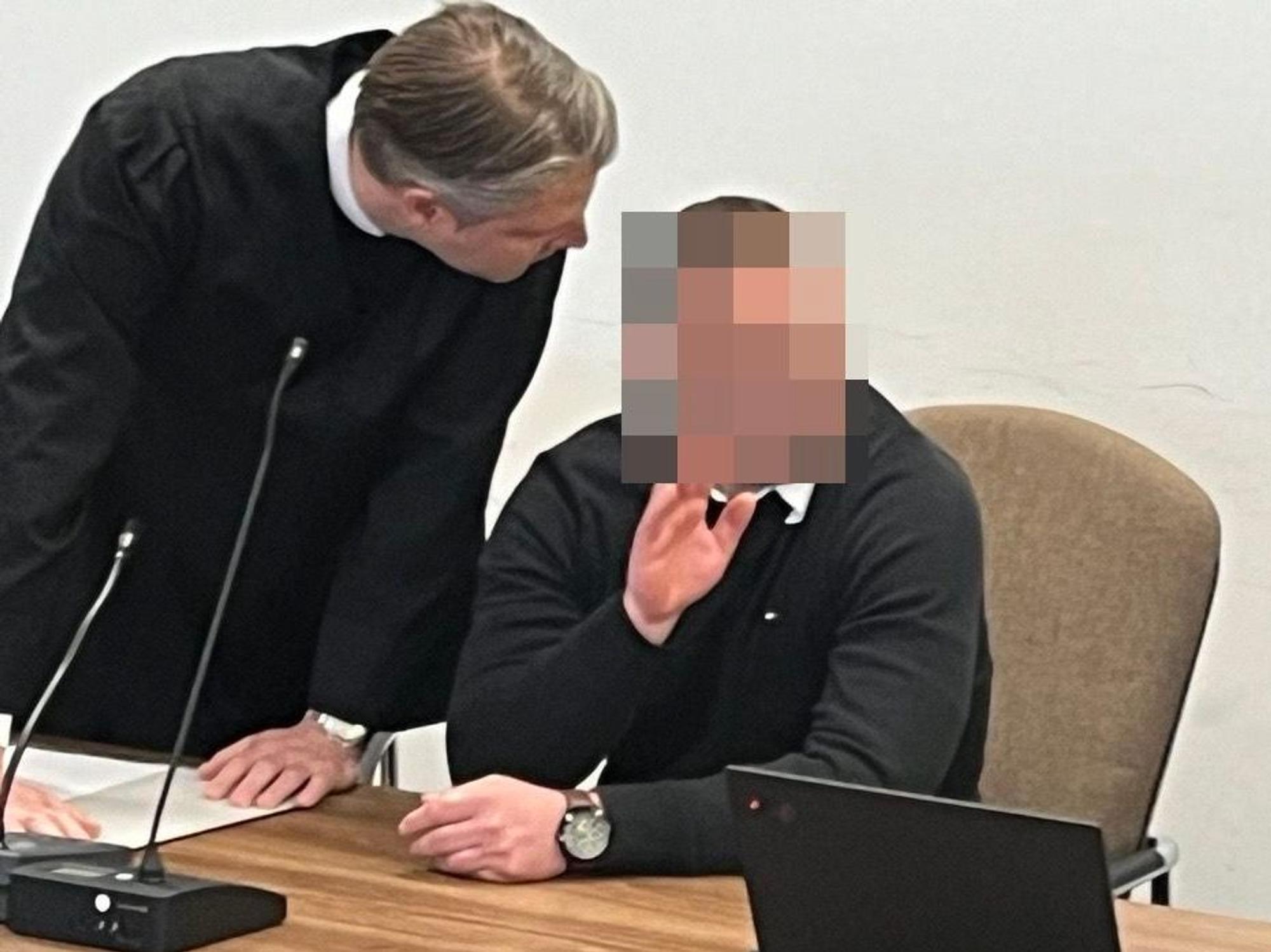 Ein Mann sitzt auf der Anklagebank und winkt, sein Anwalt beugt sich zu ihm runter.